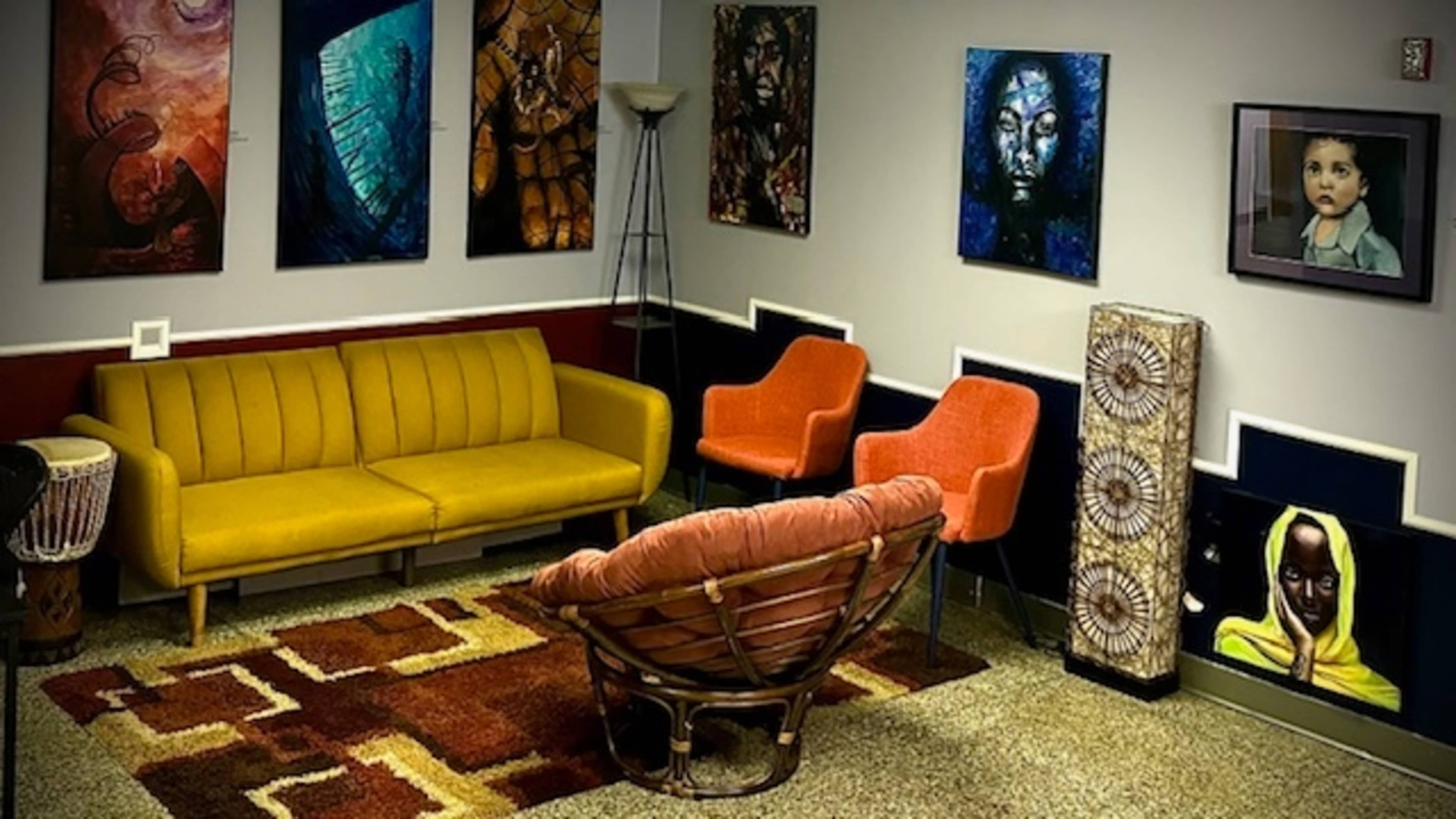 The image shows a modern waiting area with a yellow sofa, two orange chairs, various artworks on the walls, and a patterned rug on the floor.
