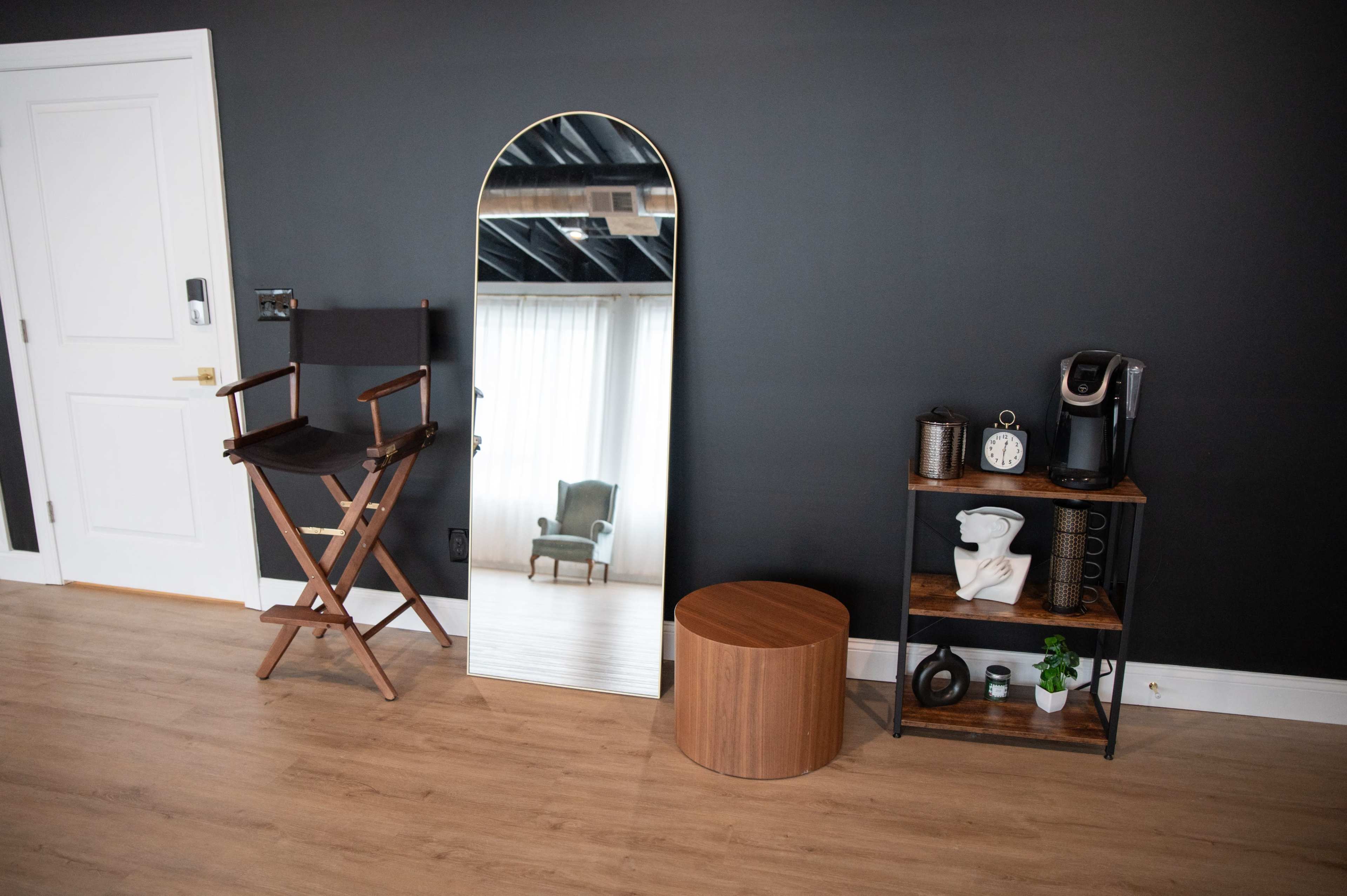A room with a large arched mirror, a wooden circular stool, a black director's chair, and a small shelving unit with a coffee maker, decorative items, and a plant.
