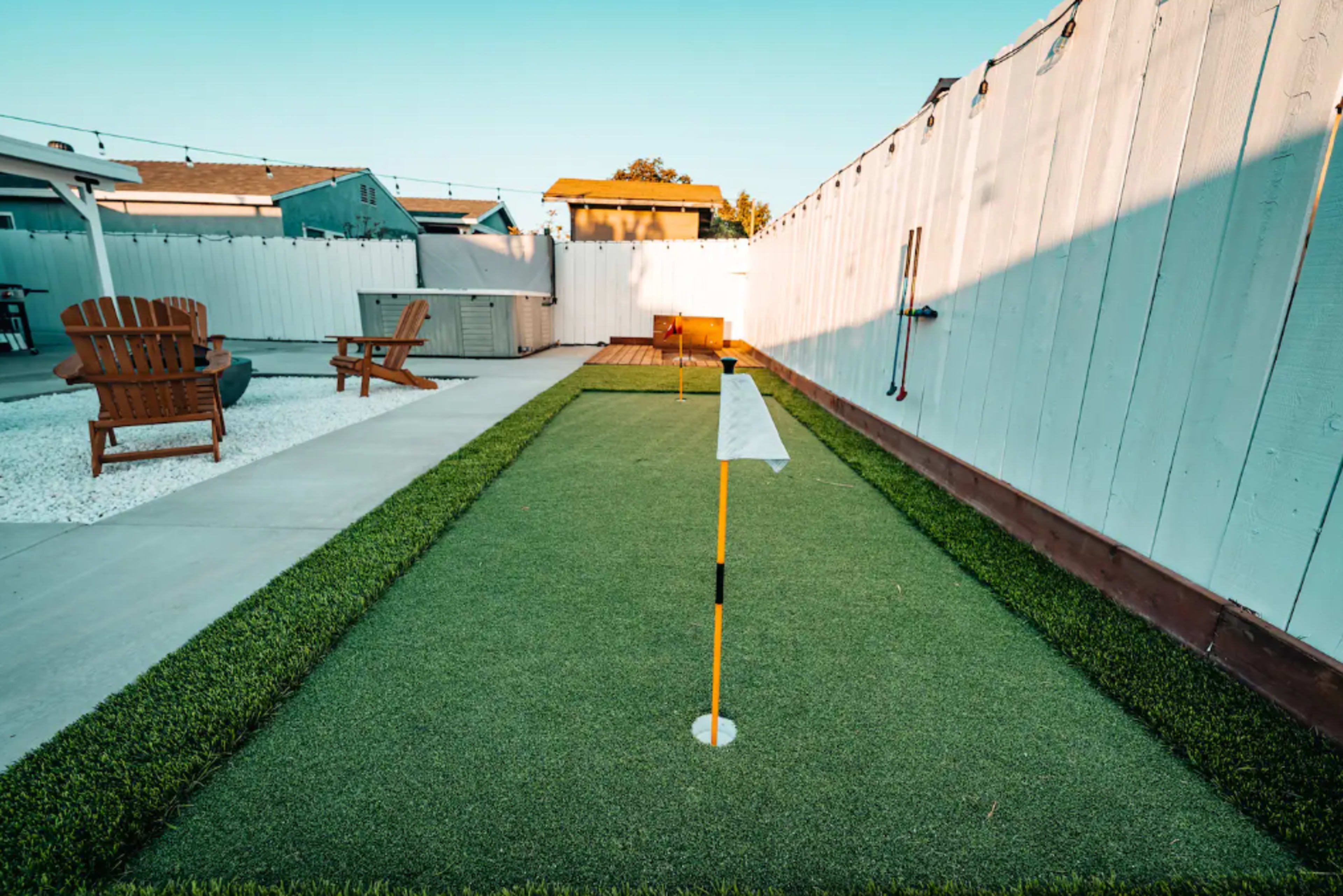 A backyard putting green bordered by artificial grass, with a flagstick and hole at one end, surrounded by a white fence and patio furniture.