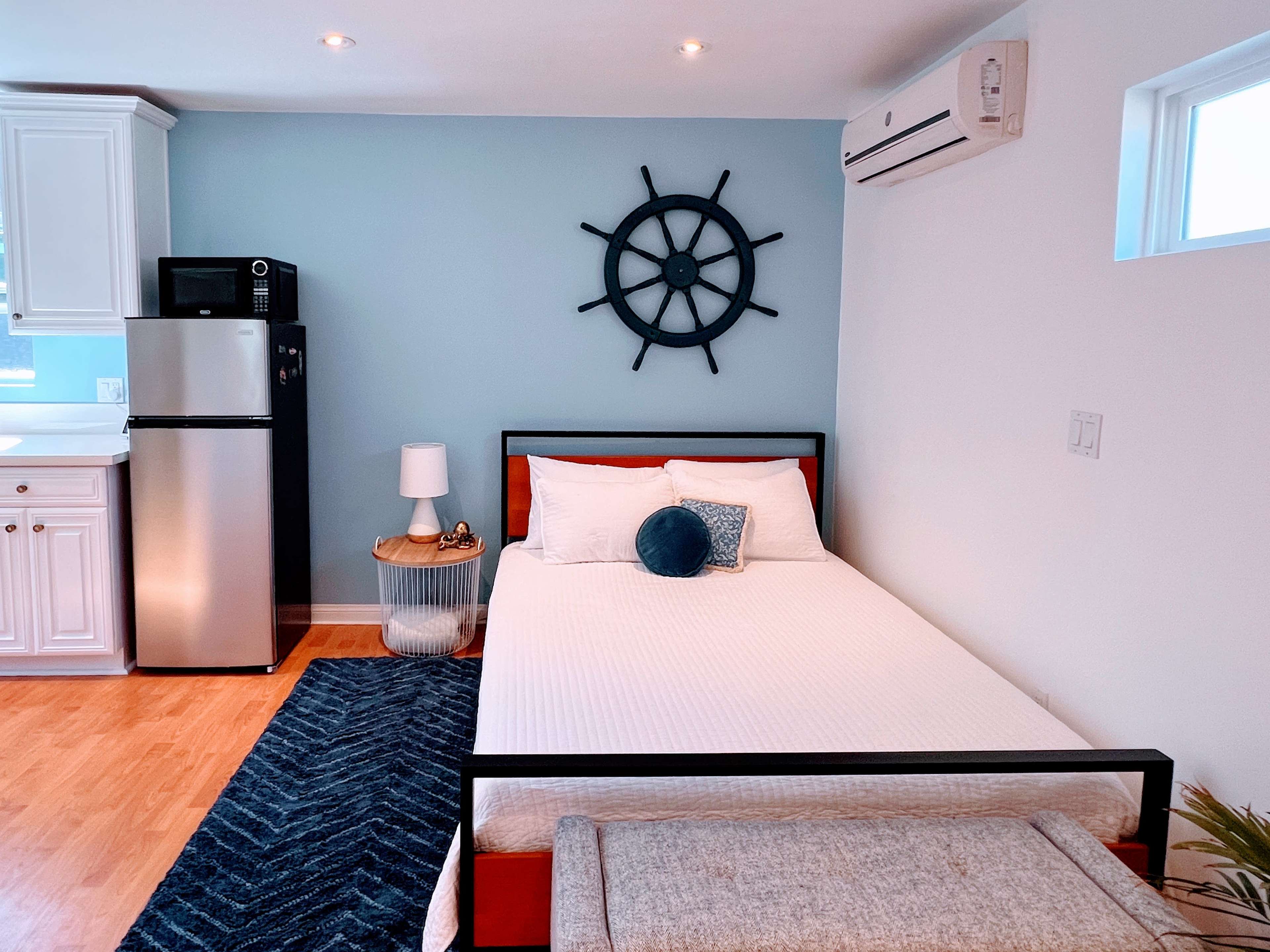 A compact bedroom featuring a bed, a bedside table with a lamp, a wall-mounted ship's wheel, a mini-fridge, and a small kitchenette area.