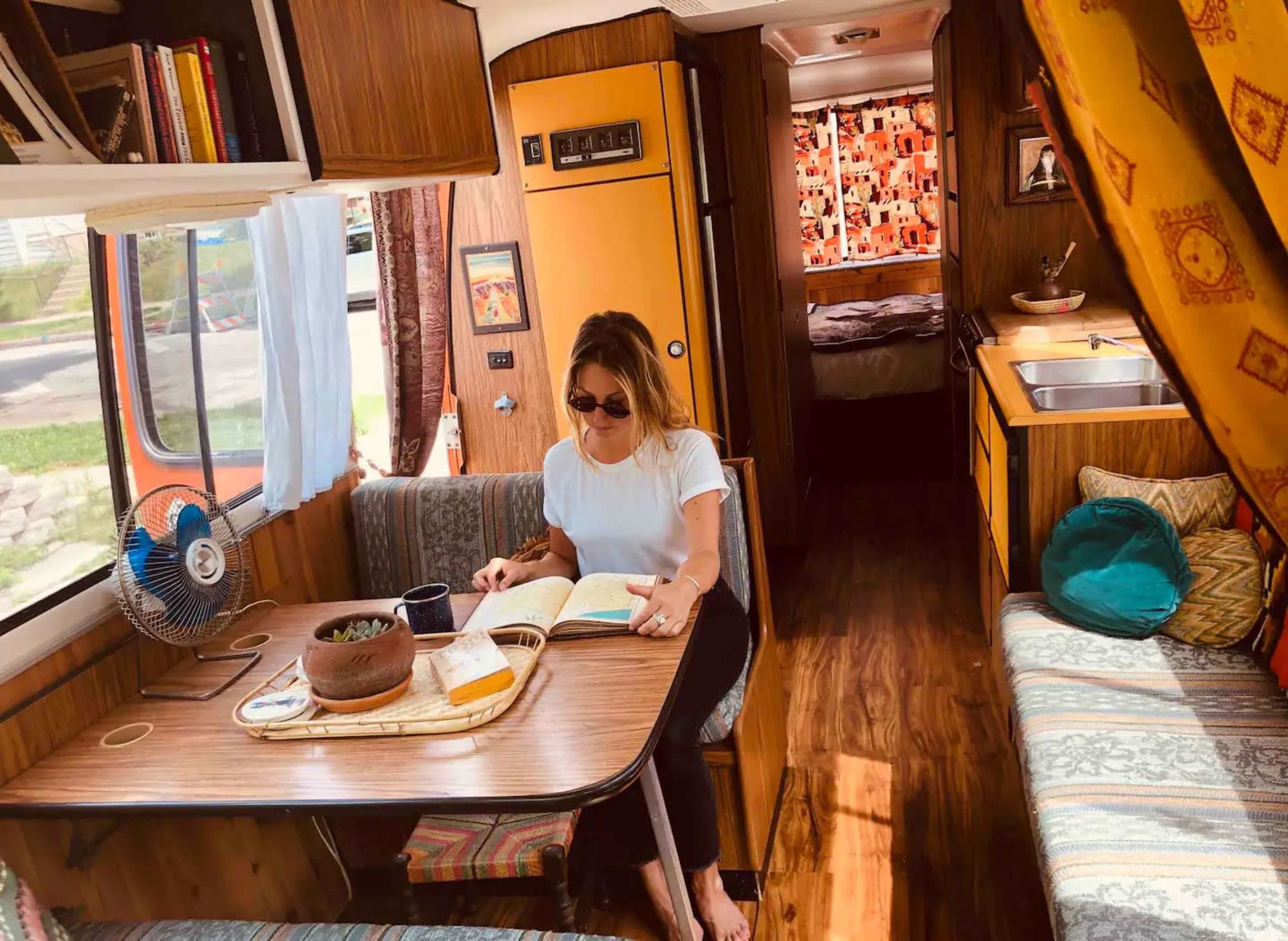 A woman sits at a table reading a book inside a vintage camper, surrounded by wooden paneling and colorful cushions.