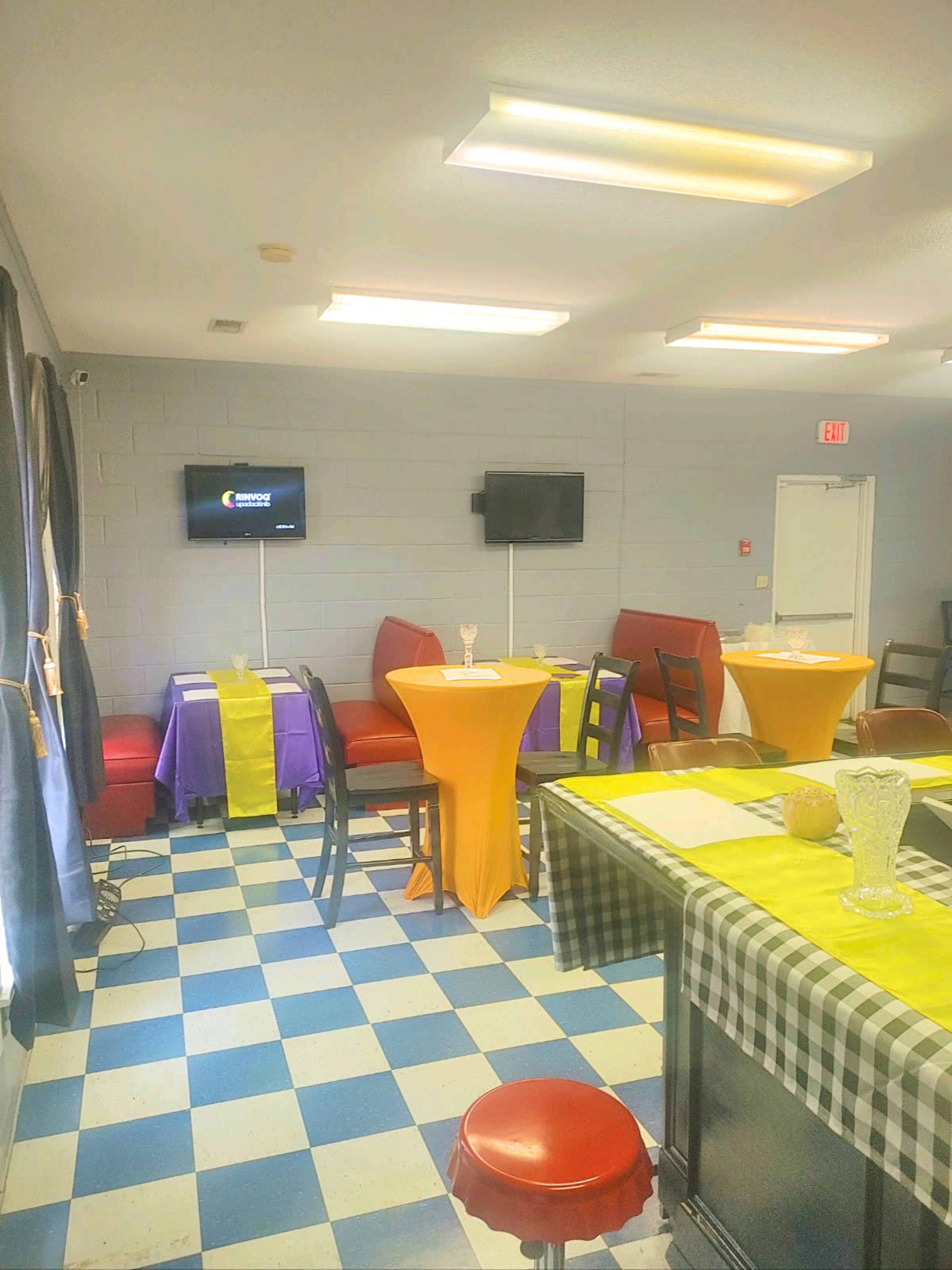A small dining area with colorful tables, red booths, wall-mounted TVs, and a checkered countertop.