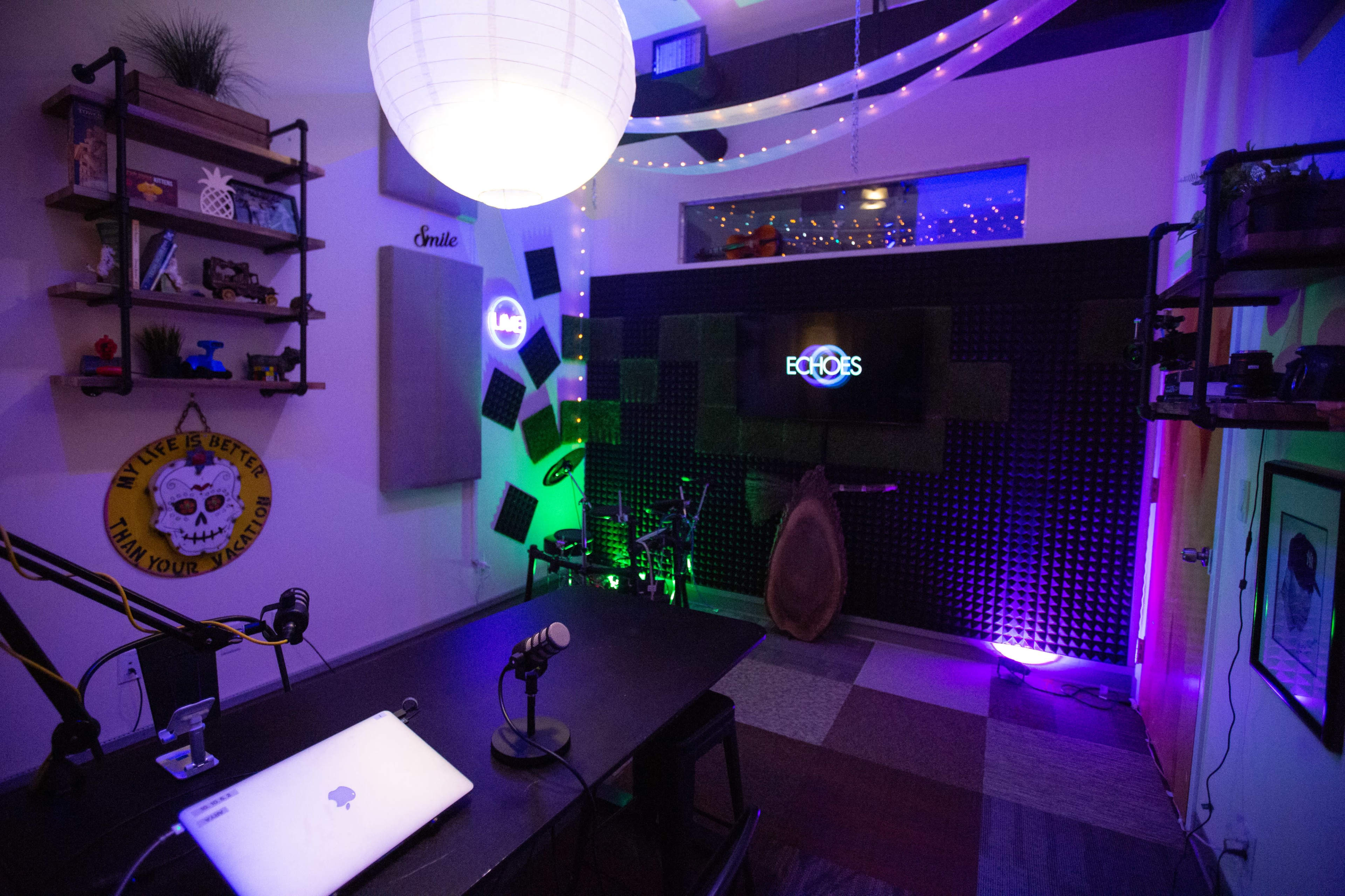 A modern recording studio featuring a table with microphones, a laptop, and colored ambient lighting, along with soundproof panels on the walls.