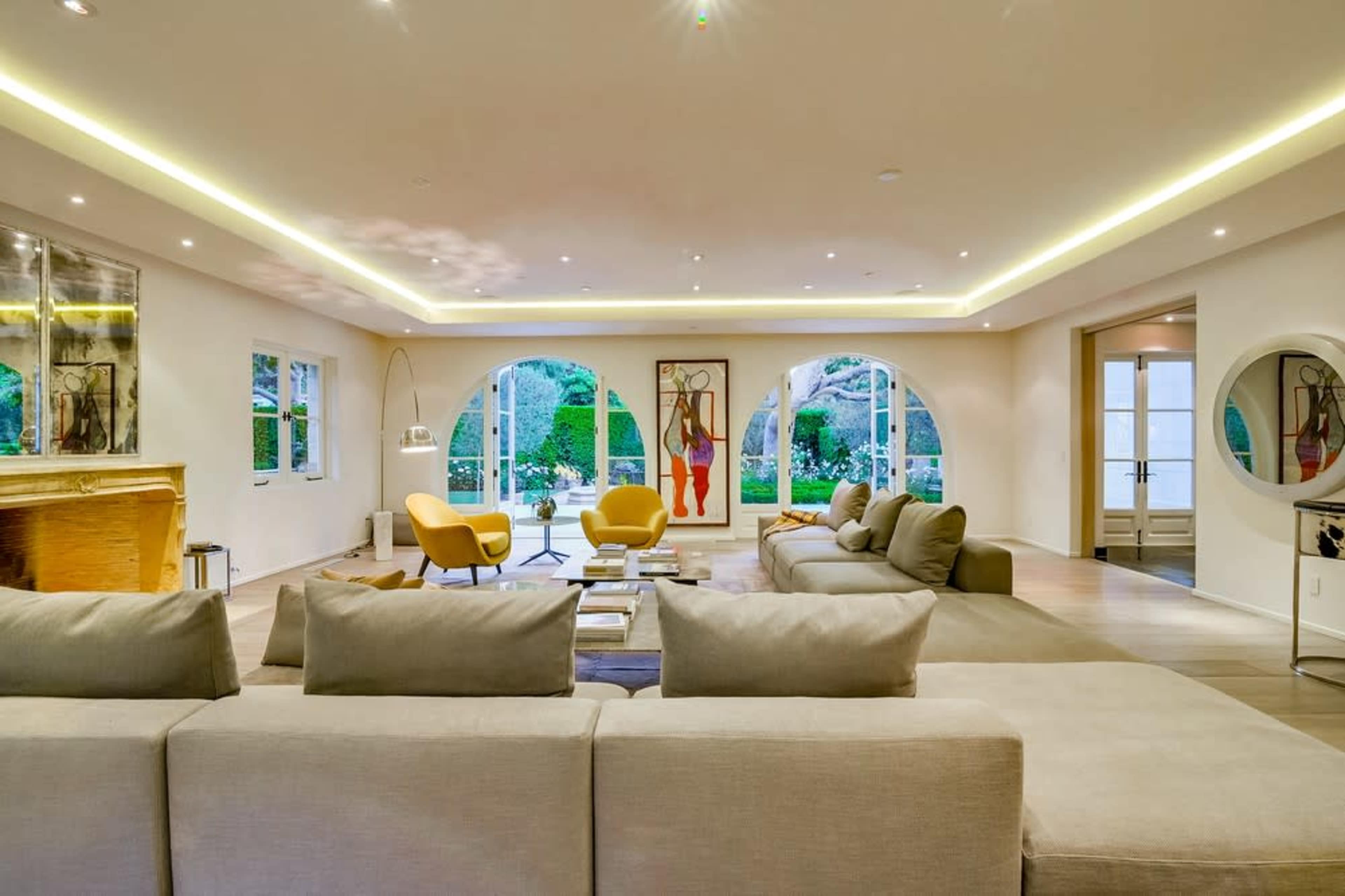 A spacious living room features a sectional sofa, two yellow armchairs, and large round windows overlooking a garden.