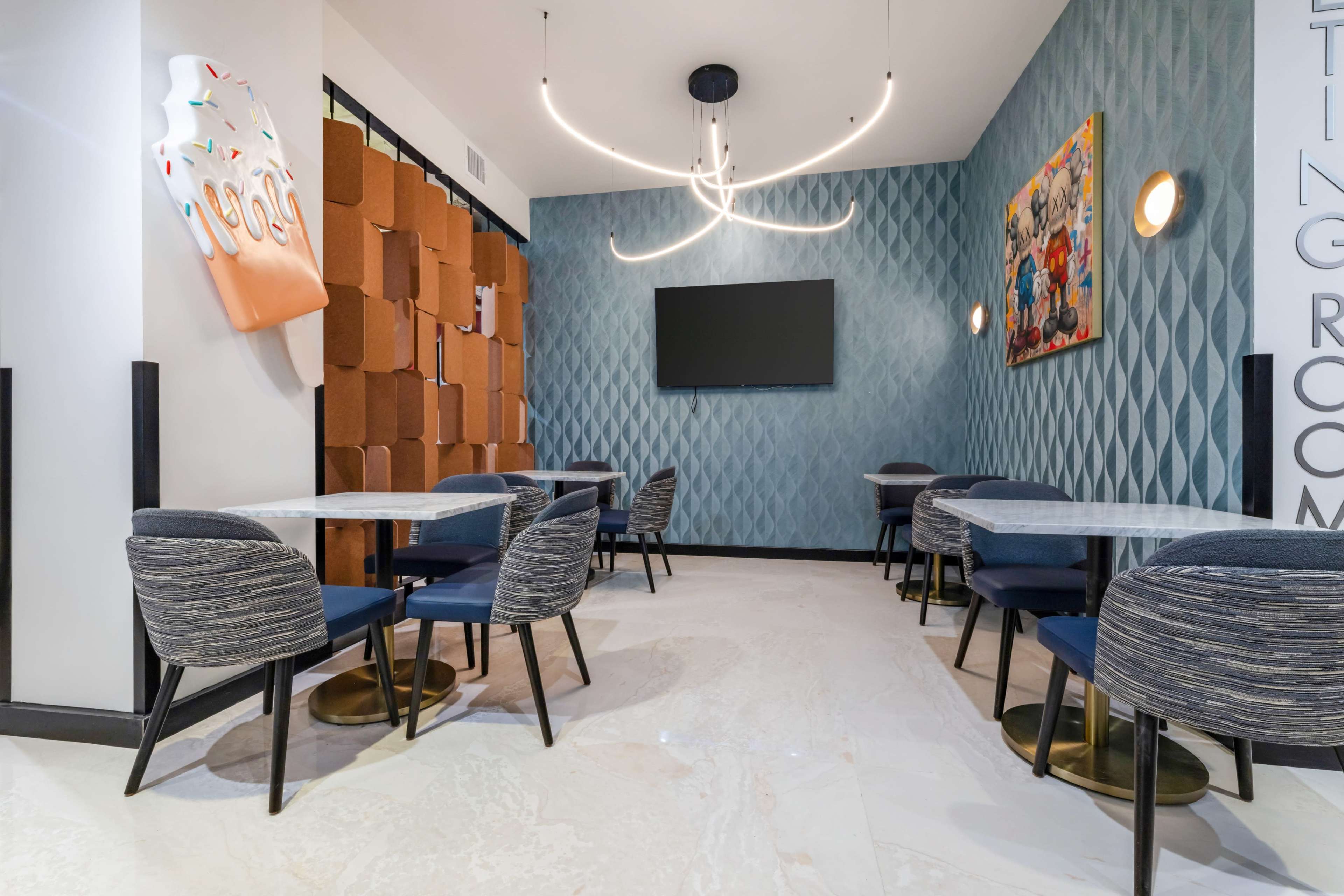 The image shows a modern dining area featuring several round tables with blue upholstered chairs, a wall adorned with decorative panels, and a television mounted on a wall.