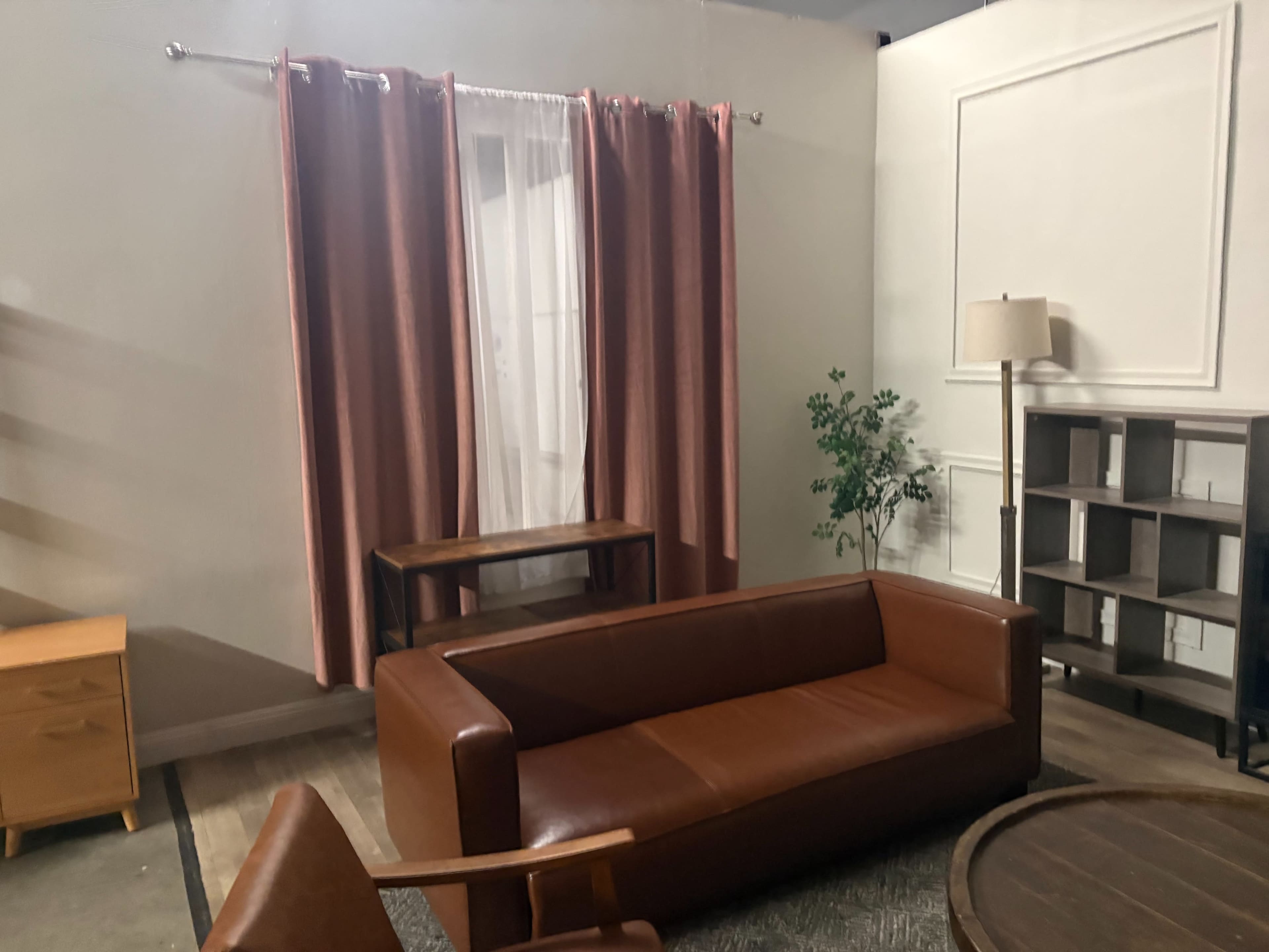 The image shows a neatly arranged living room featuring a brown leather sofa, a wooden coffee table, a floor lamp, and curtains, with a potted plant in the corner.