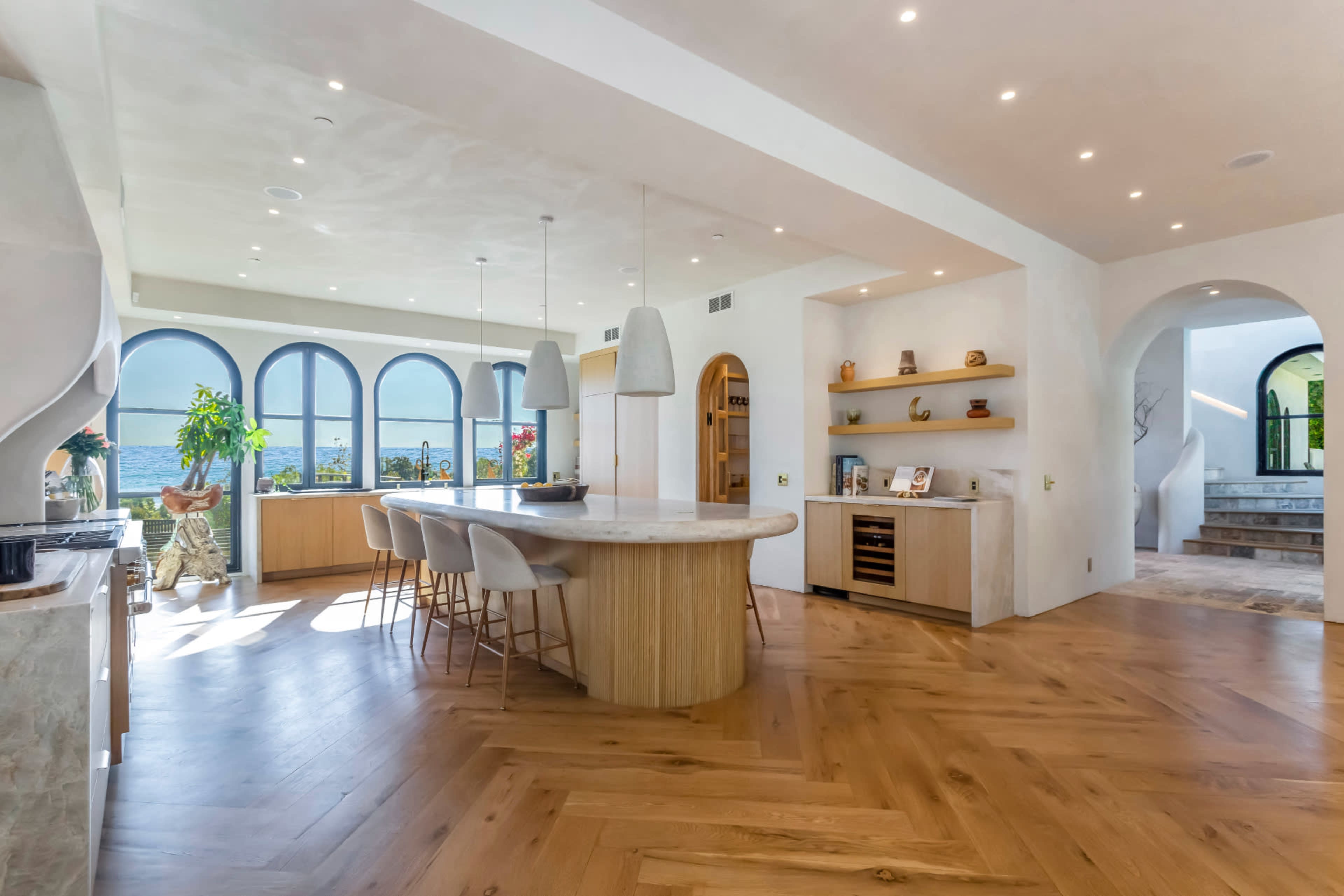 The image shows a modern kitchen and dining area with a large island, bar seating, and windows overlooking a body of water.