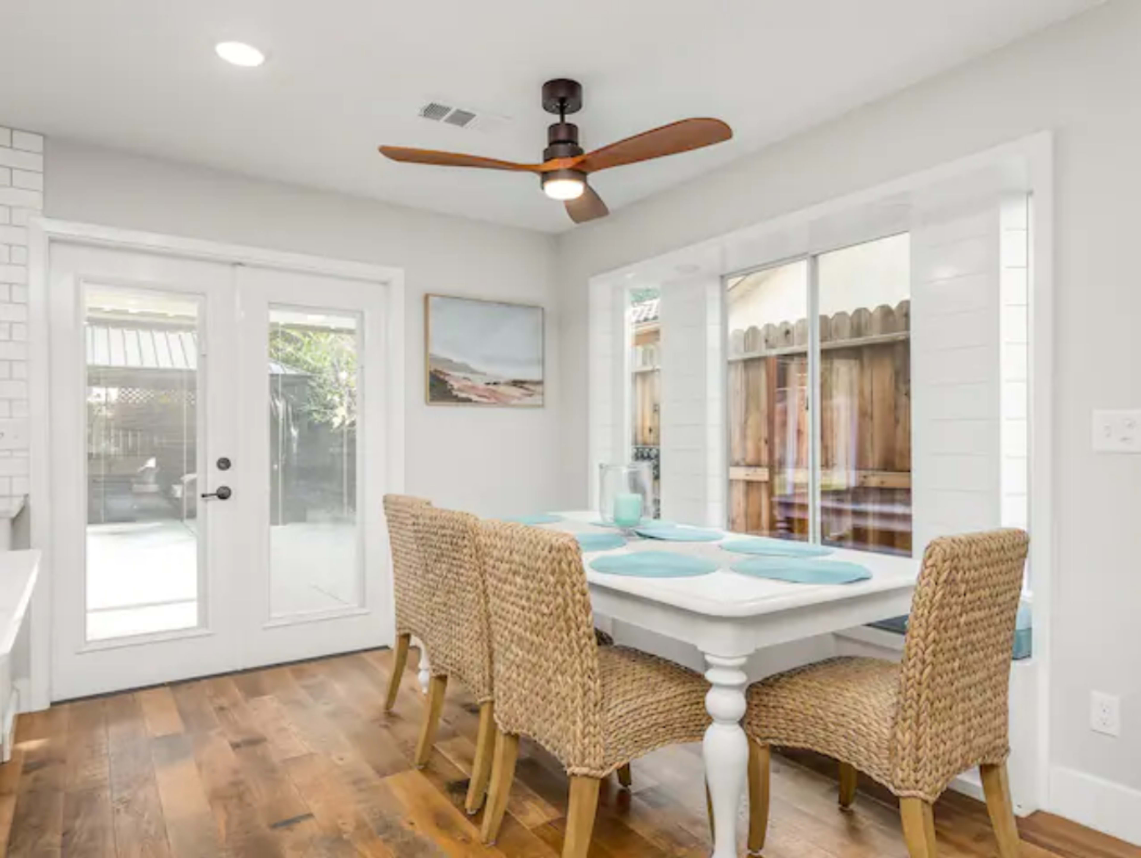 A dining area features a white table surrounded by four wicker chairs, with a ceiling fan and large windows overlooking a wooden fence.