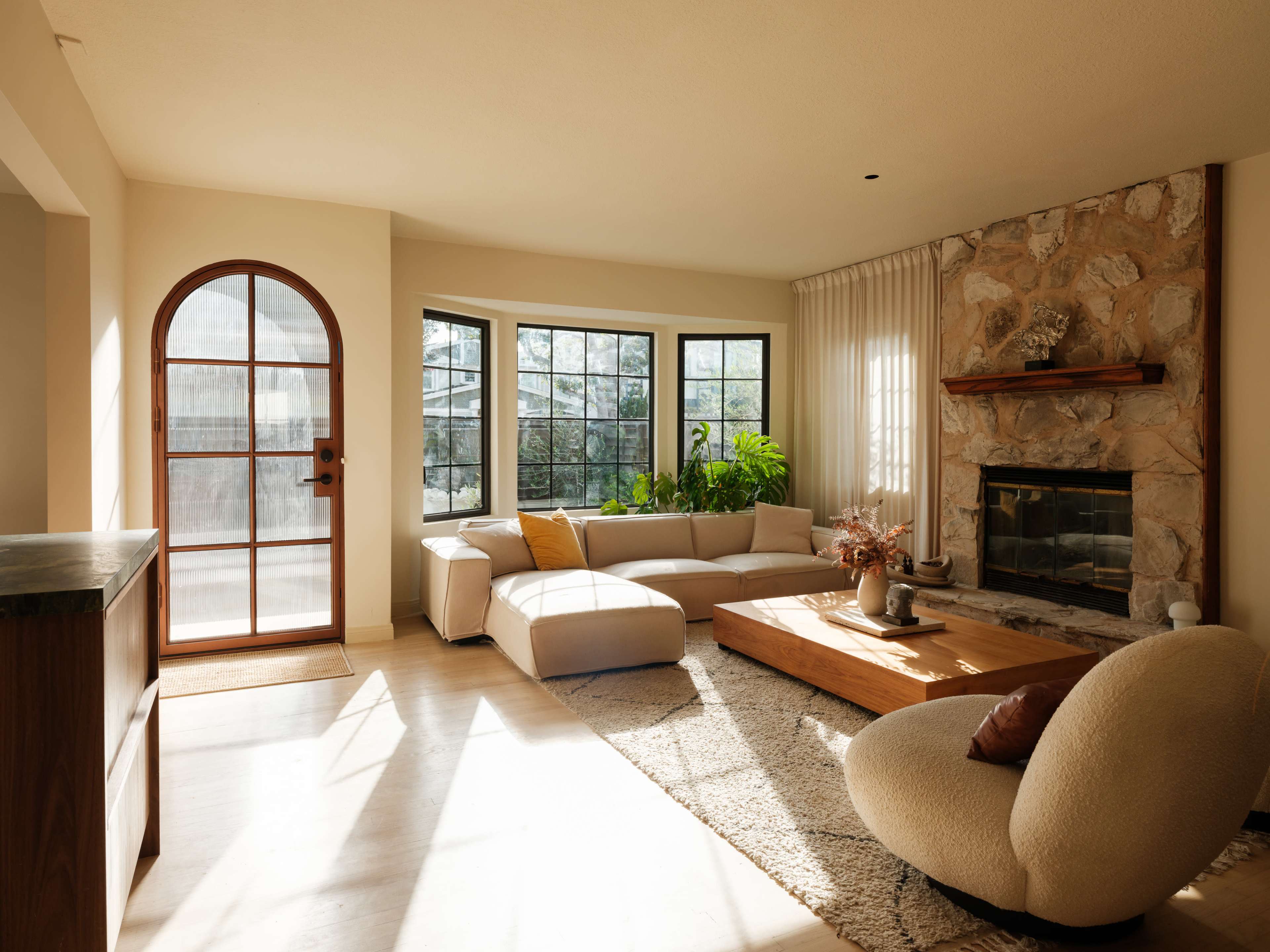 The living room features a light-colored sectional sofa, a wooden coffee table, and a stone fireplace, with large windows allowing natural light to fill the space.