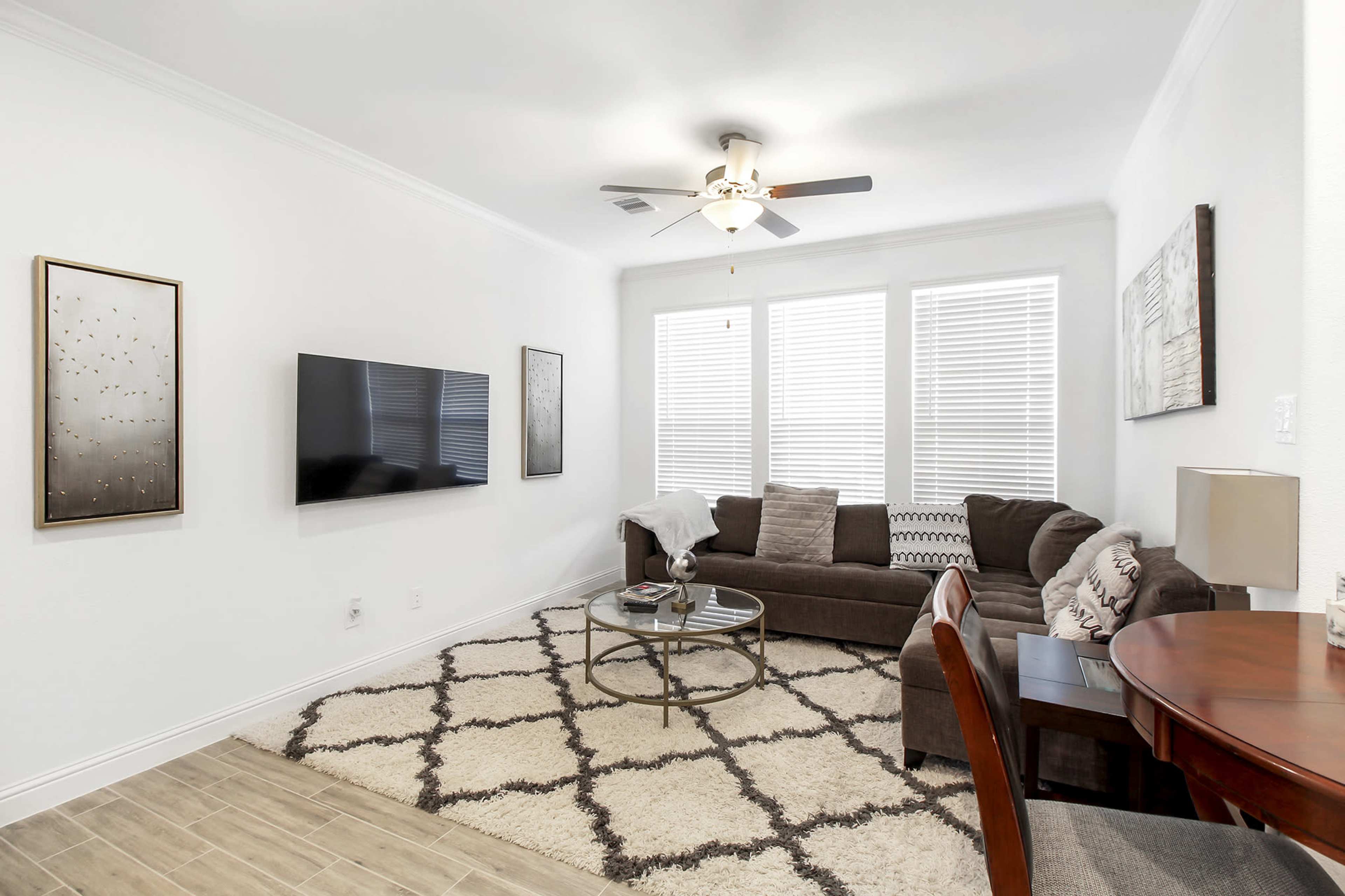 The room features a modern living space with a sectional sofa, a coffee table, a television mounted on the wall, and large windows allowing natural light.
