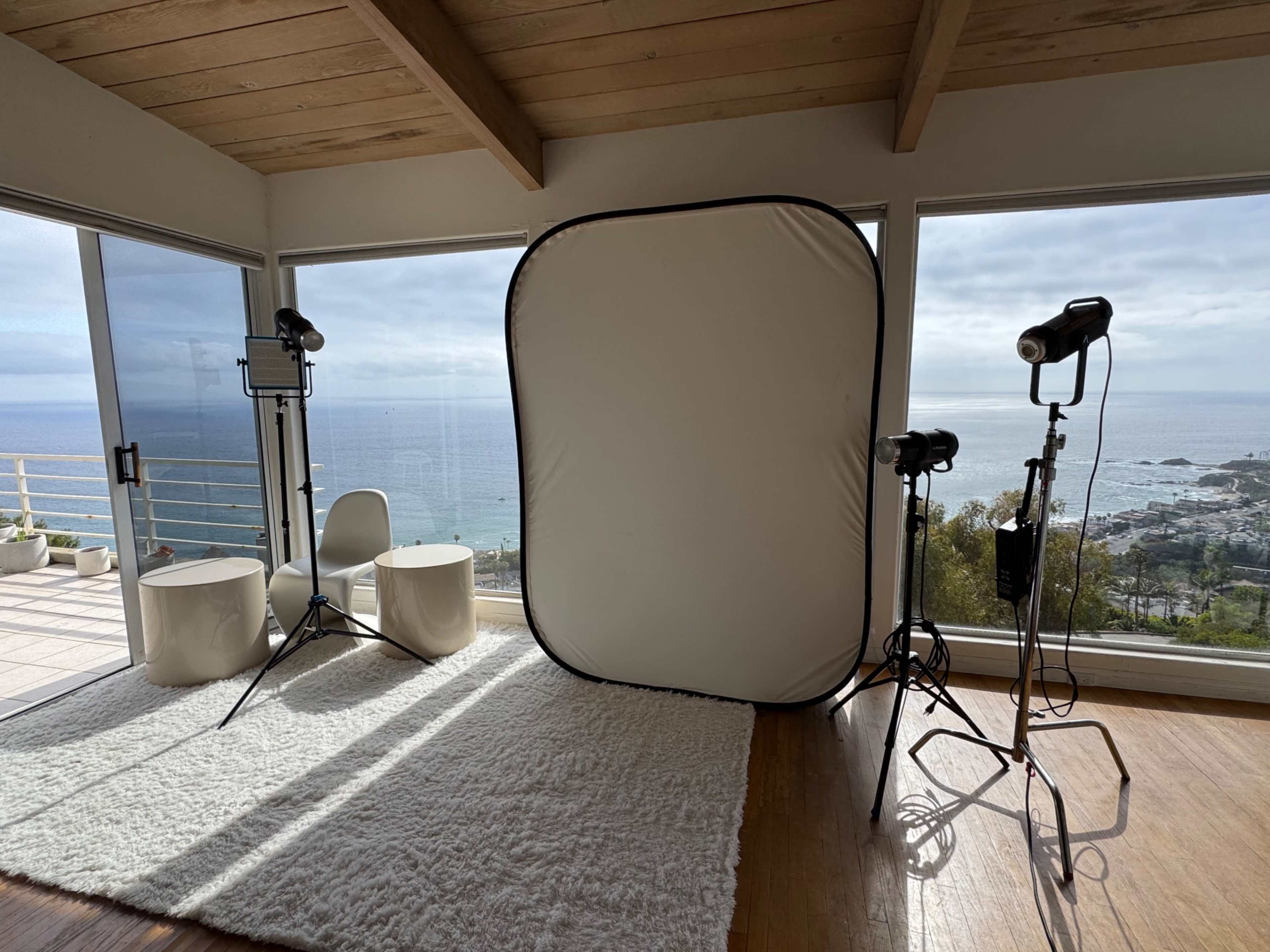 A photography setup is arranged in a room with large windows overlooking the ocean, featuring a backdrop and lighting equipment on stands.