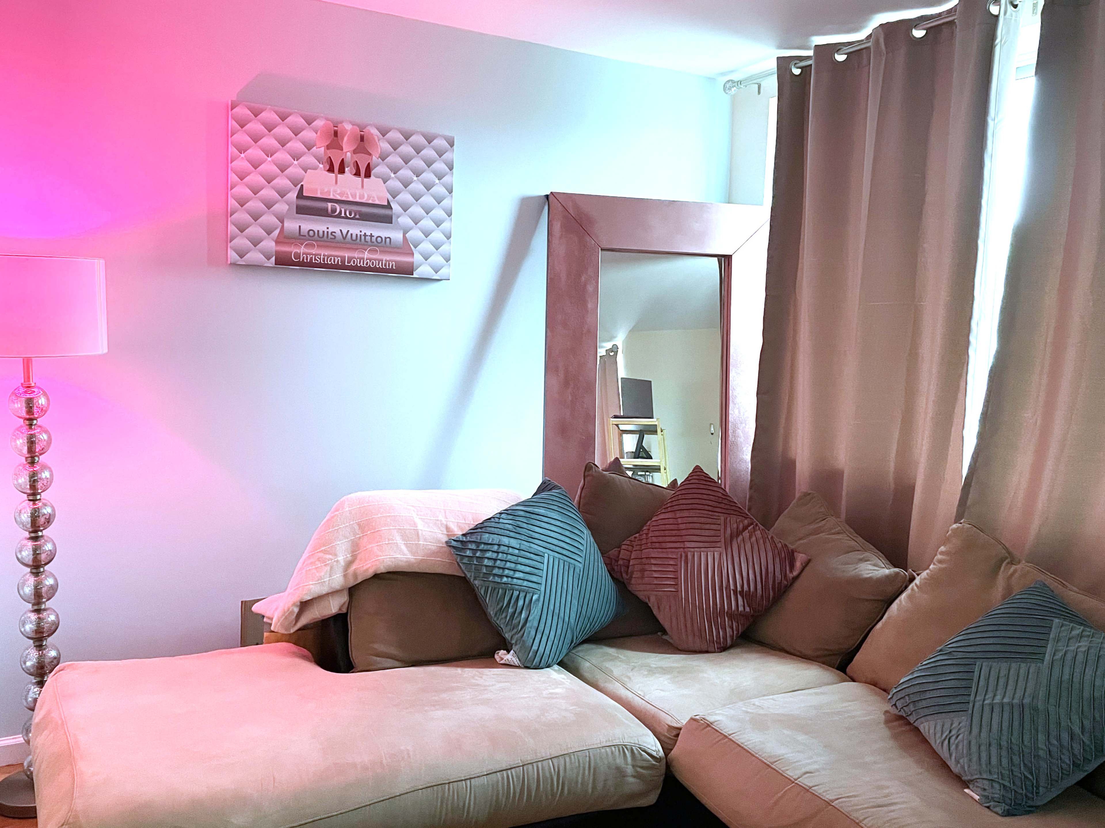 A living room corner with a beige sectional sofa, a large mirror, and a decorative wall art piece featuring fashion branding.