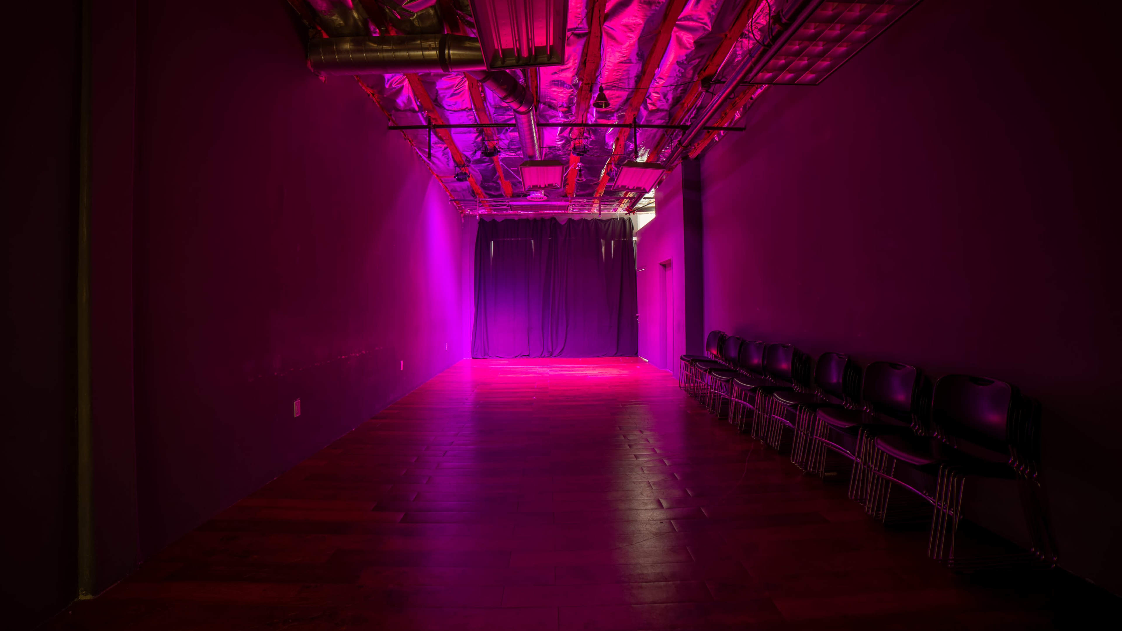 A long, narrow room with a polished wooden floor, illuminated by purple lights, features black chairs neatly arranged against one wall.