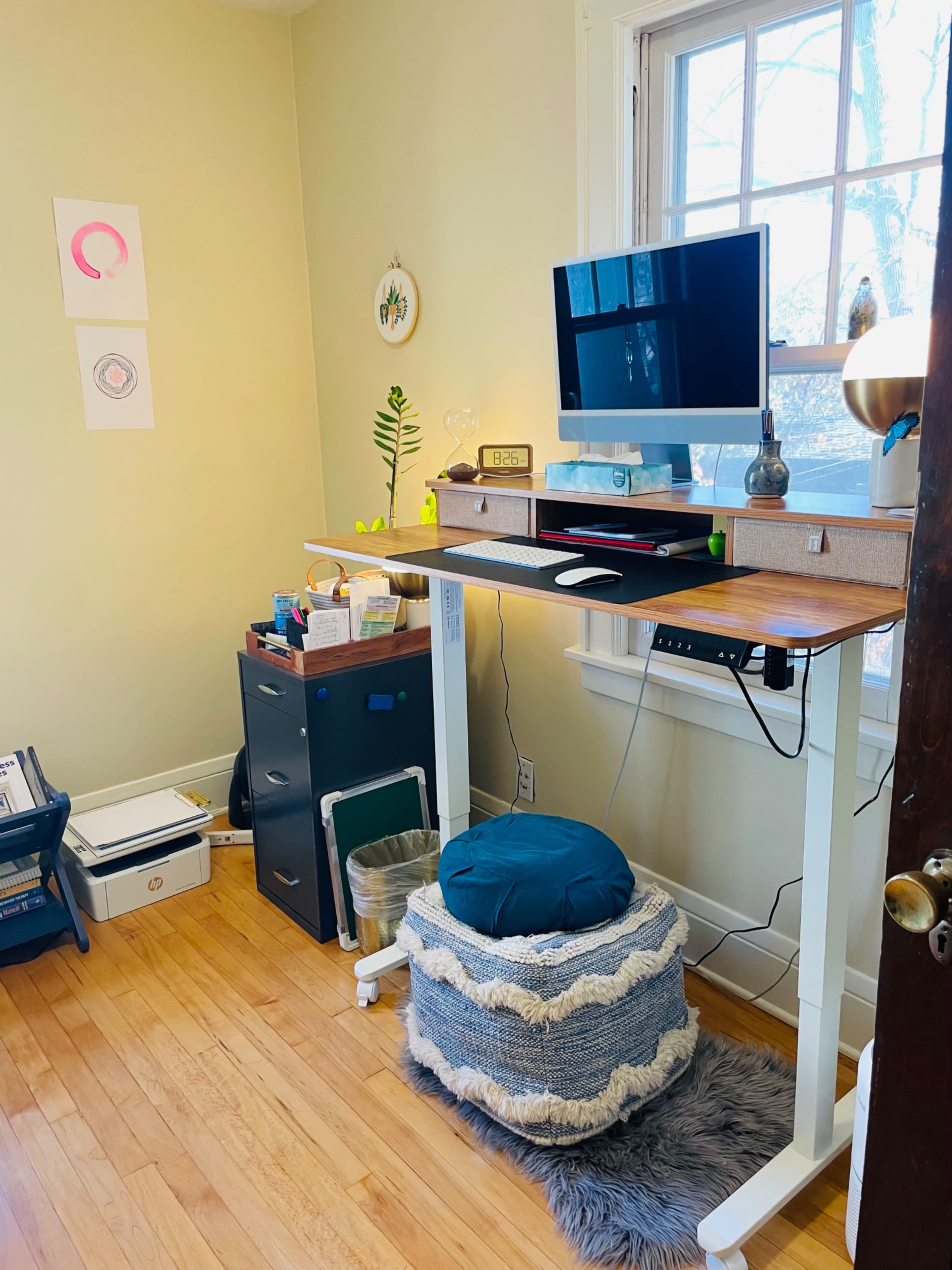 A standing desk with a monitor and lamp is positioned against a window in a room featuring a storage cabinet and decorative items on the walls.