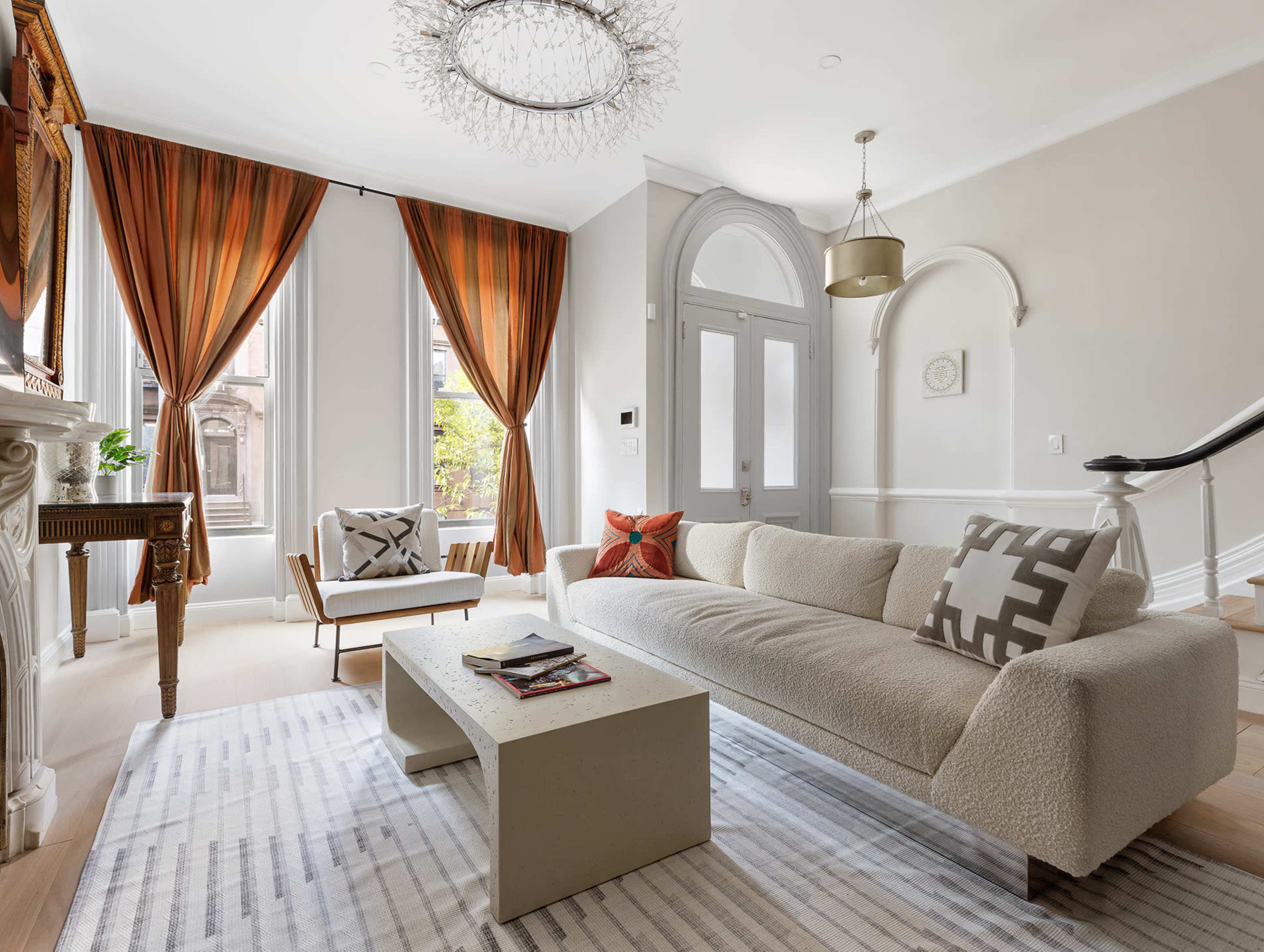 The image shows a modern living room featuring a light-colored sofa, a rectangular coffee table, and large windows dressed with orange and brown curtains.