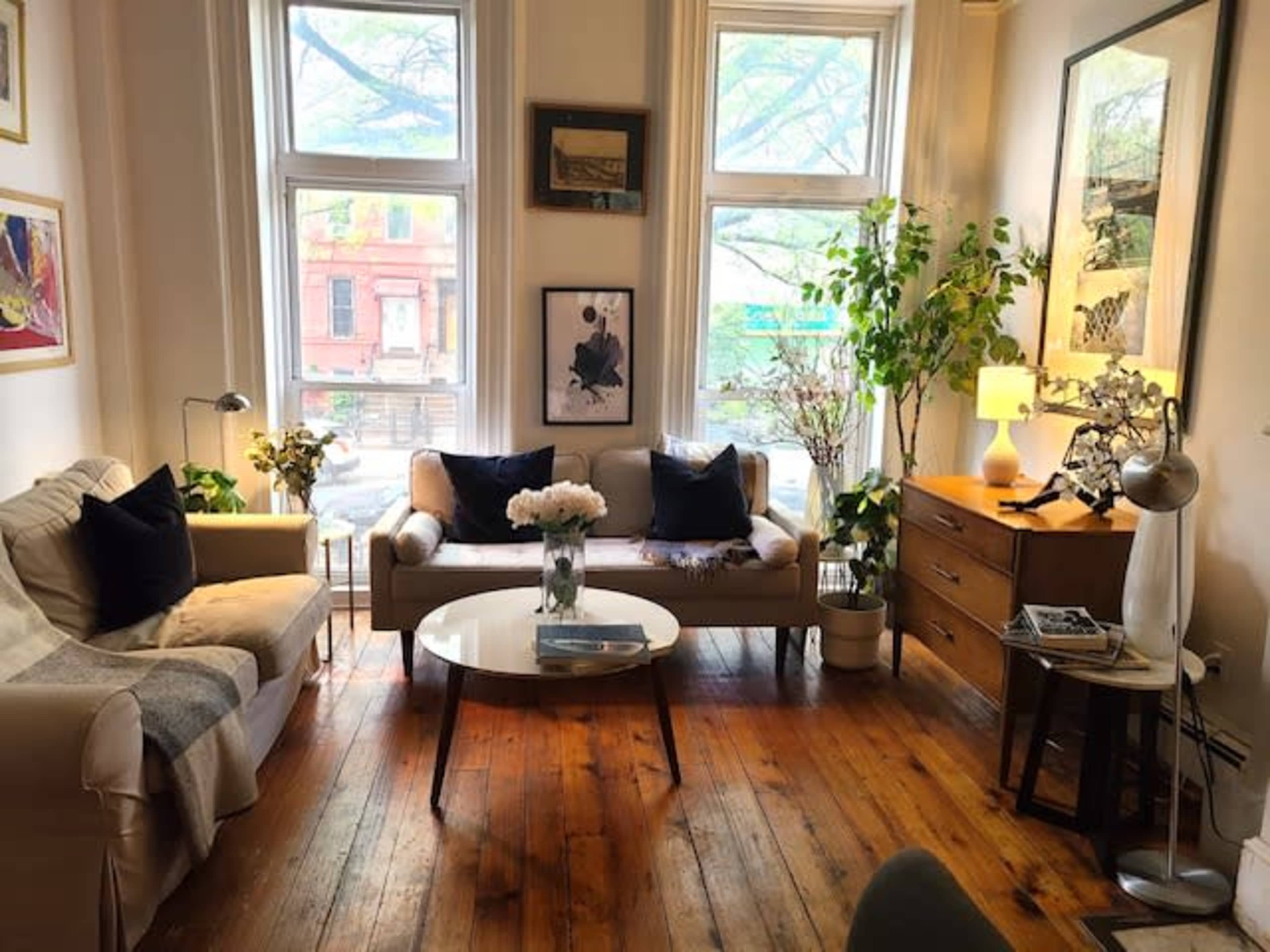 A cozy living room featuring two sofas, a round coffee table, and a wooden side table, with large windows letting in natural light and plants placed throughout the space.