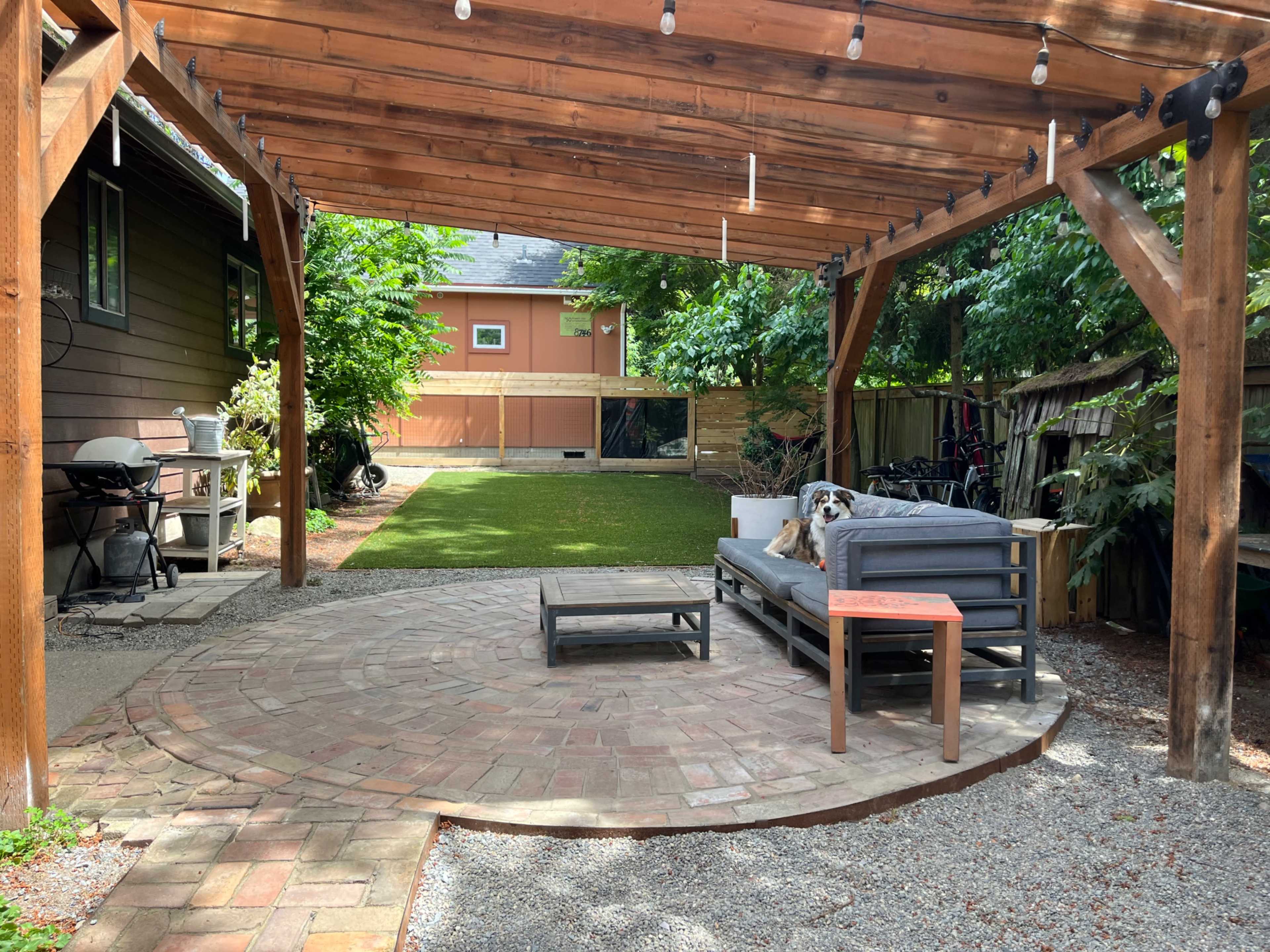 The image shows a patio area with a wooden pergola, featuring a seating arrangement, a small table, and a garden space in the background.