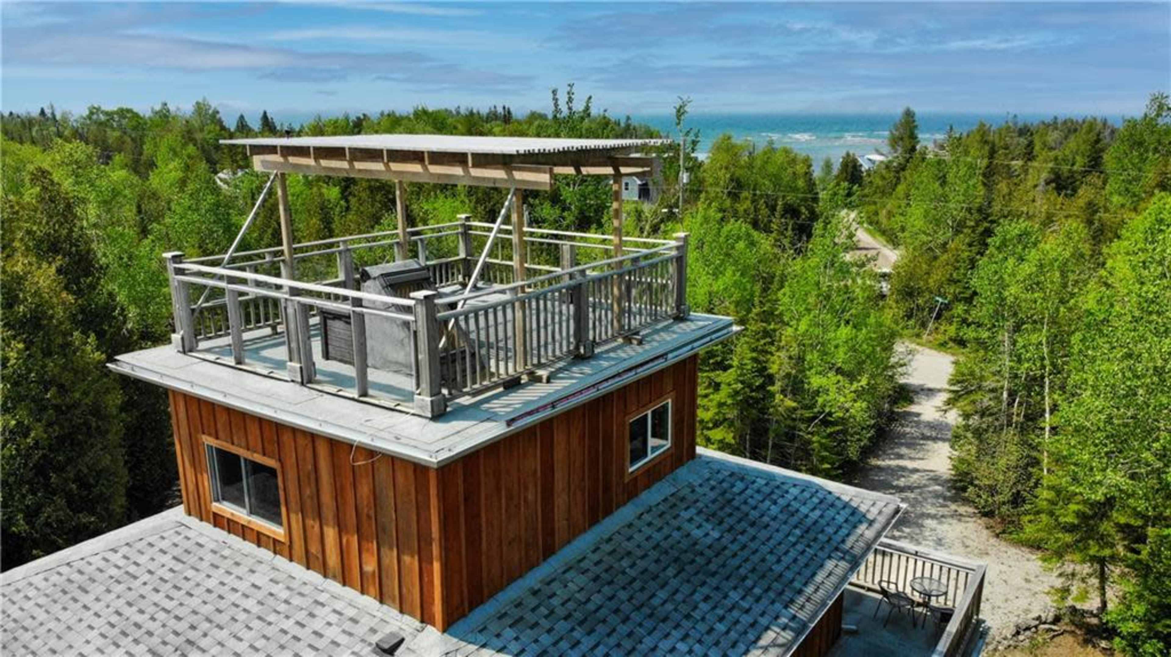 The image shows a wooden cabin with a deck on its rooftop, surrounded by dense trees and a gravel pathway leading into the distance.