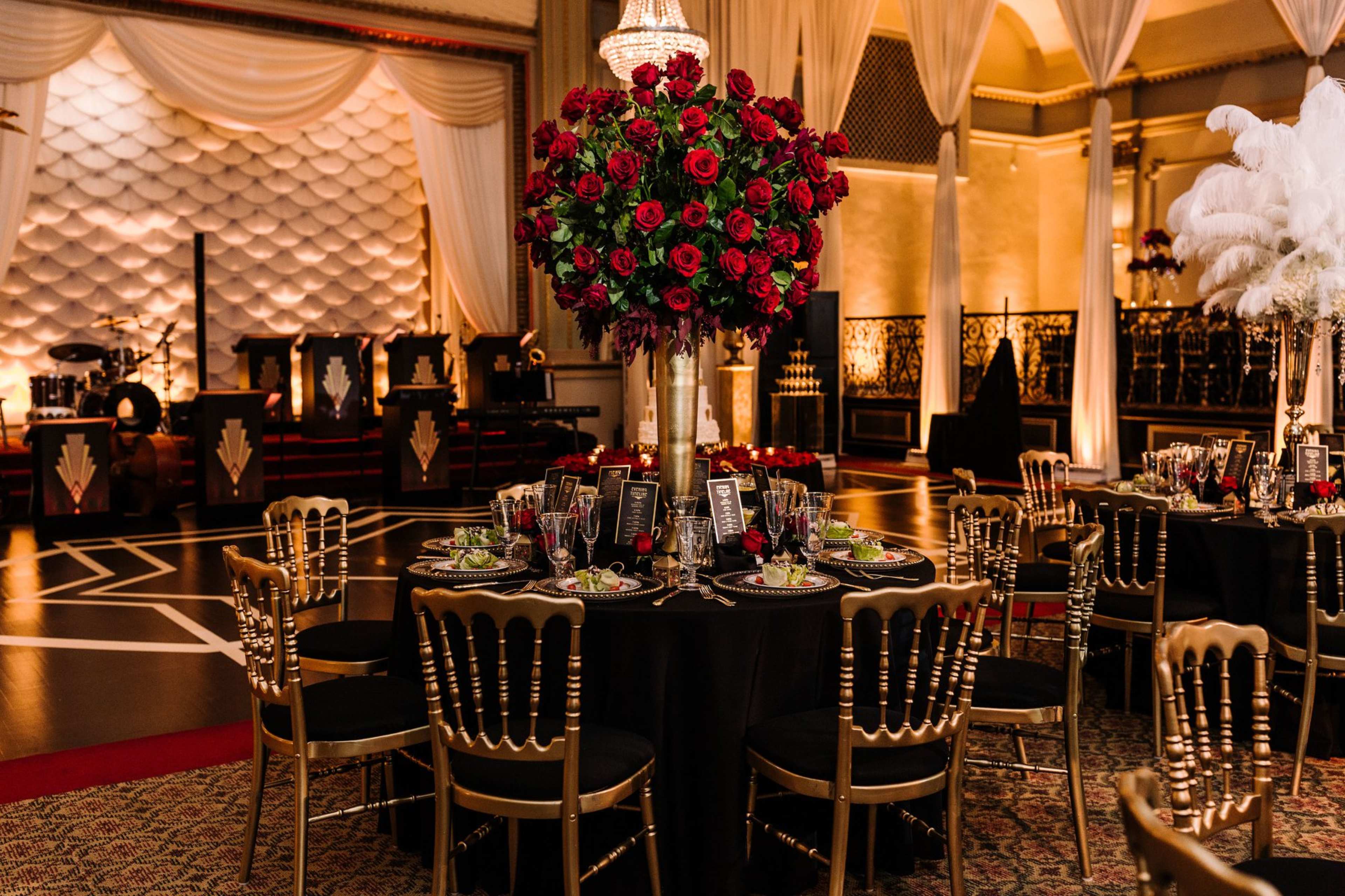 A beautifully arranged dining area features a round table with a tall floral centerpiece of red roses, surrounded by elegant gold chairs and set with black tablecloths and fine dining plates.