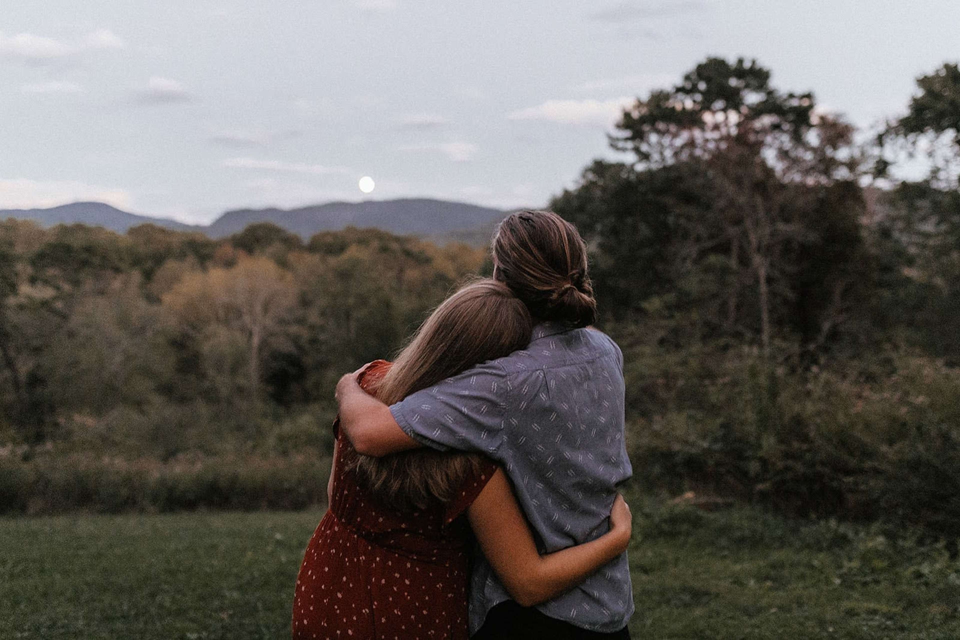 A couple stands together in an open field, embracing while facing a mountain landscape at dusk.