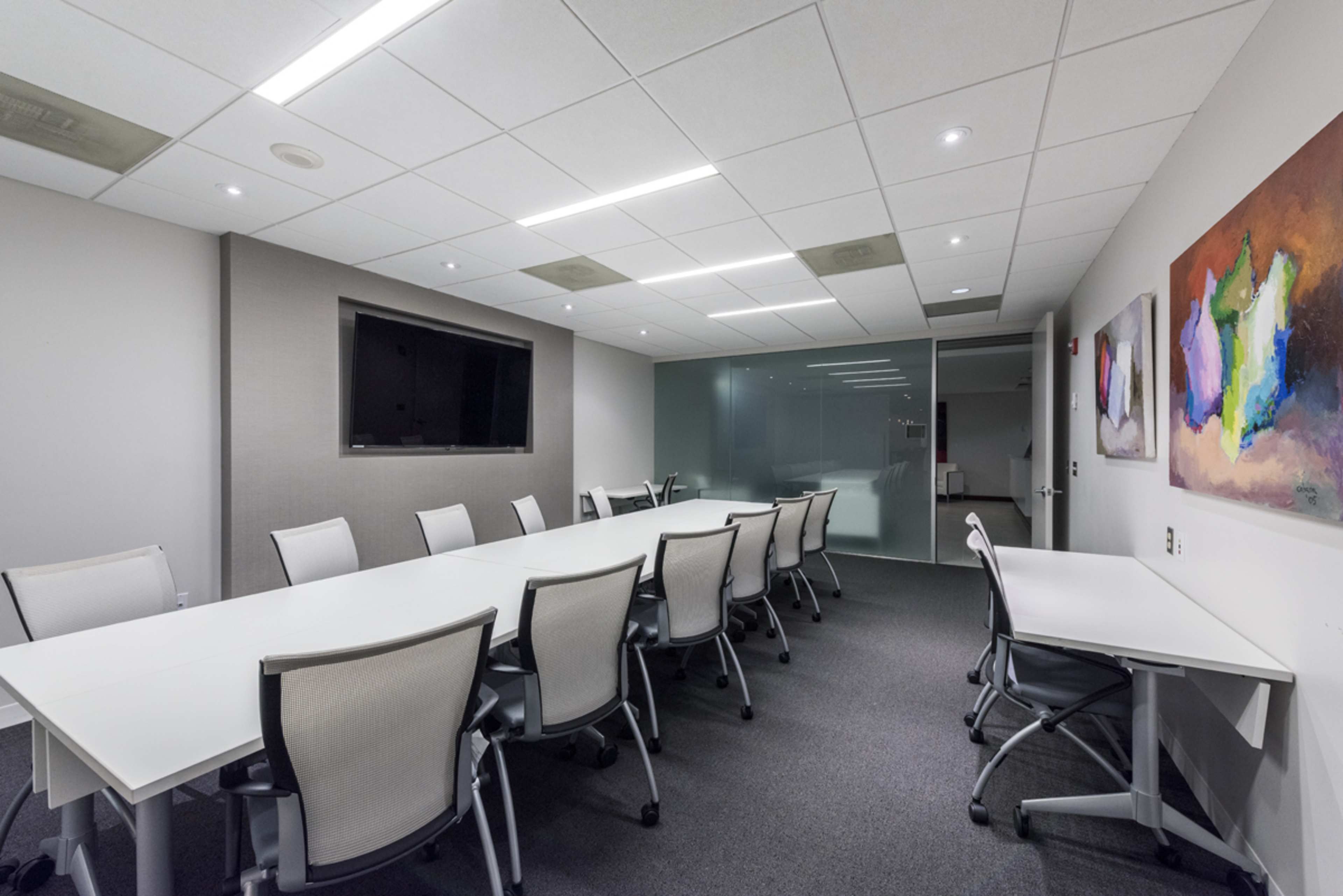 A large, empty conference room features a long white table surrounded by ergonomic chairs and a wall-mounted television.