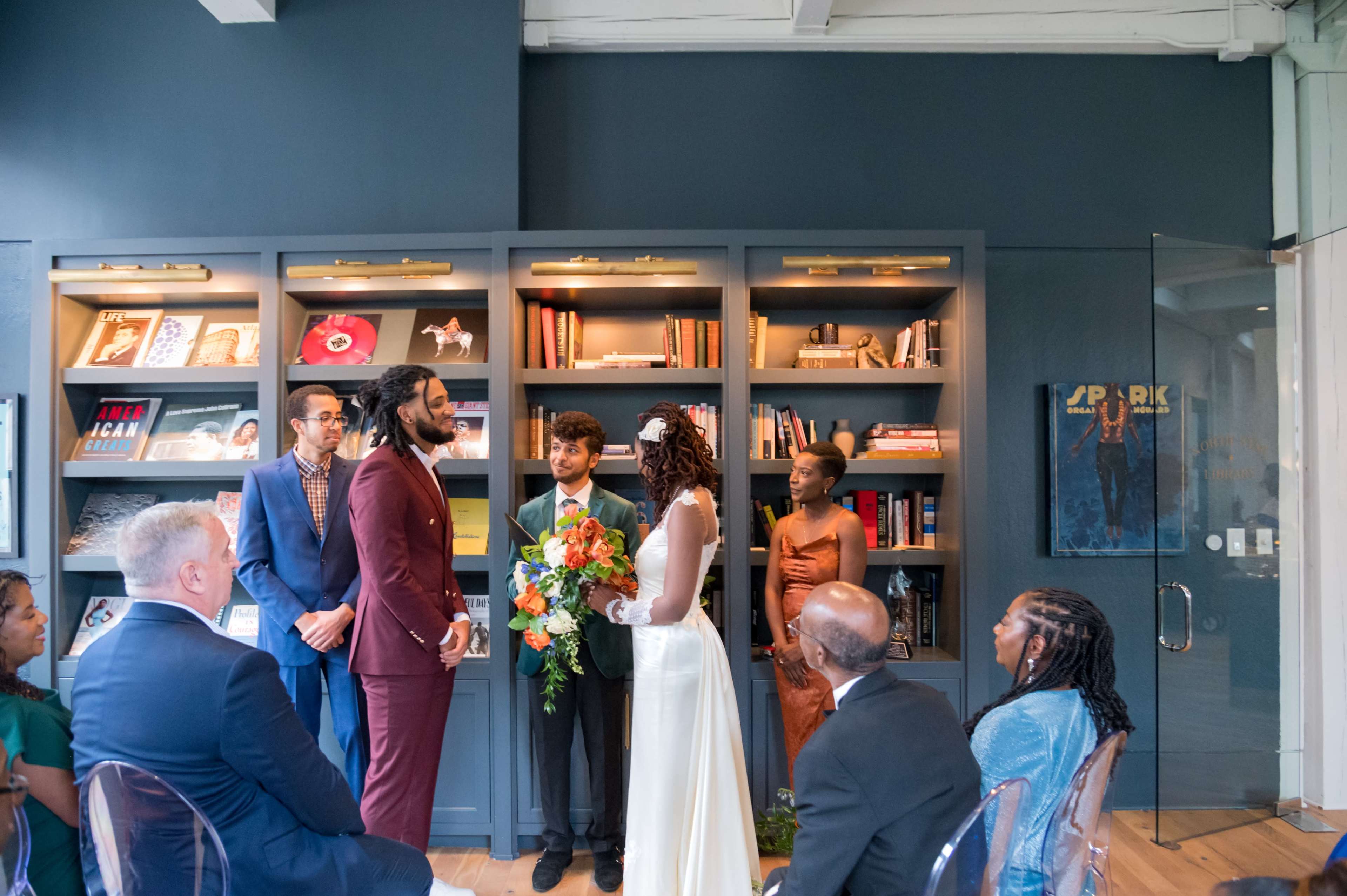 A couple stands at the front of a modern room adorned with bookshelves and artwork, exchanging vows while surrounded by their wedding party and guests.