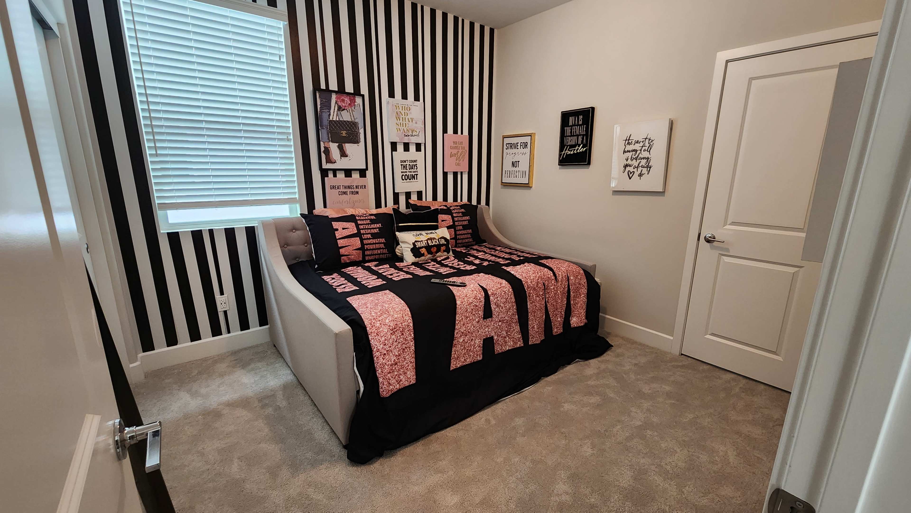 A bedroom with a bed covered by a black and pink "I AM" patterned duvet and walls featuring black and white striped wallpaper along with framed artwork.