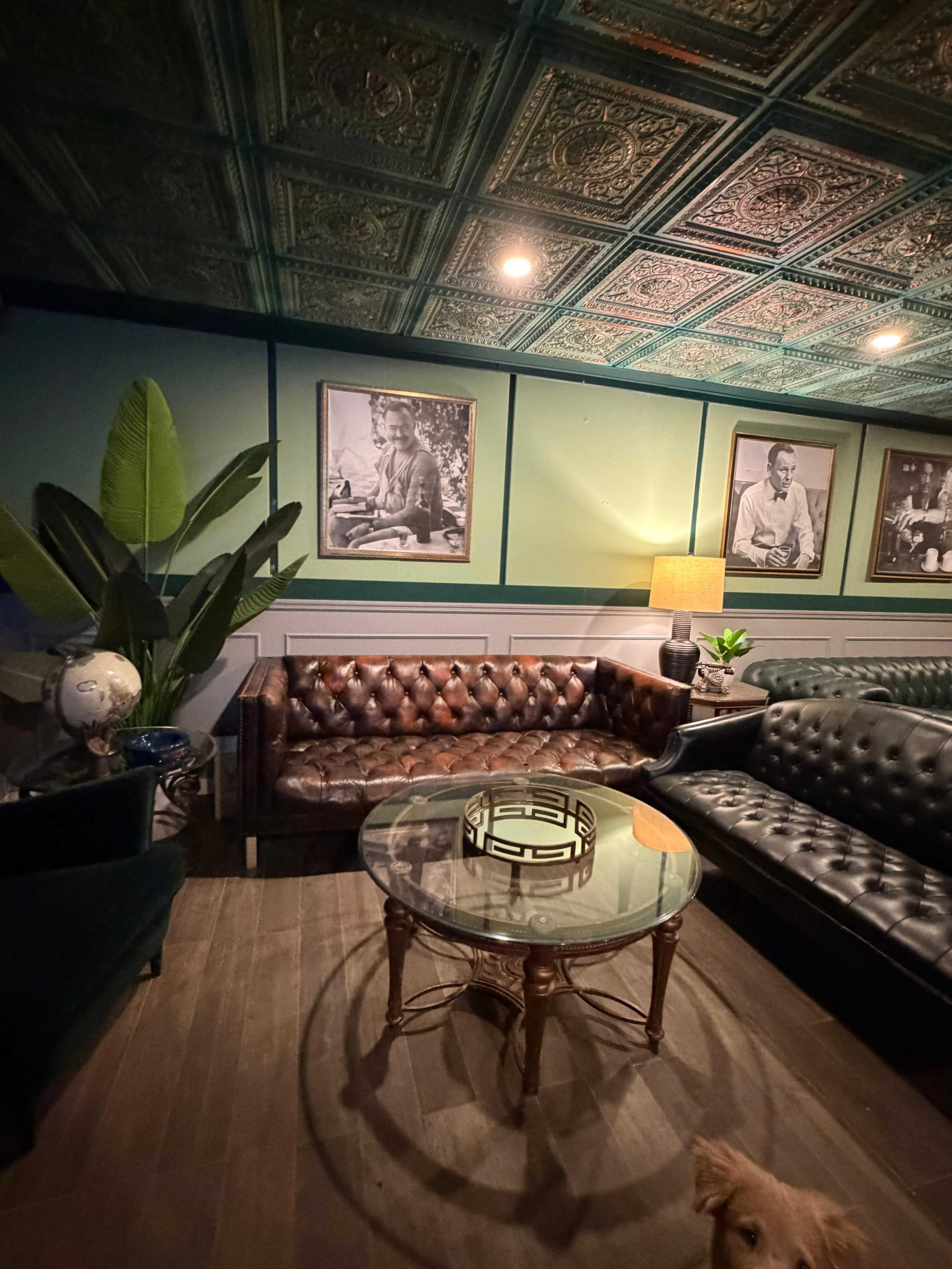 The image shows a cozy lounge area with a leather sofa, a glass coffee table, potted plants, and black and white framed photographs on the walls.