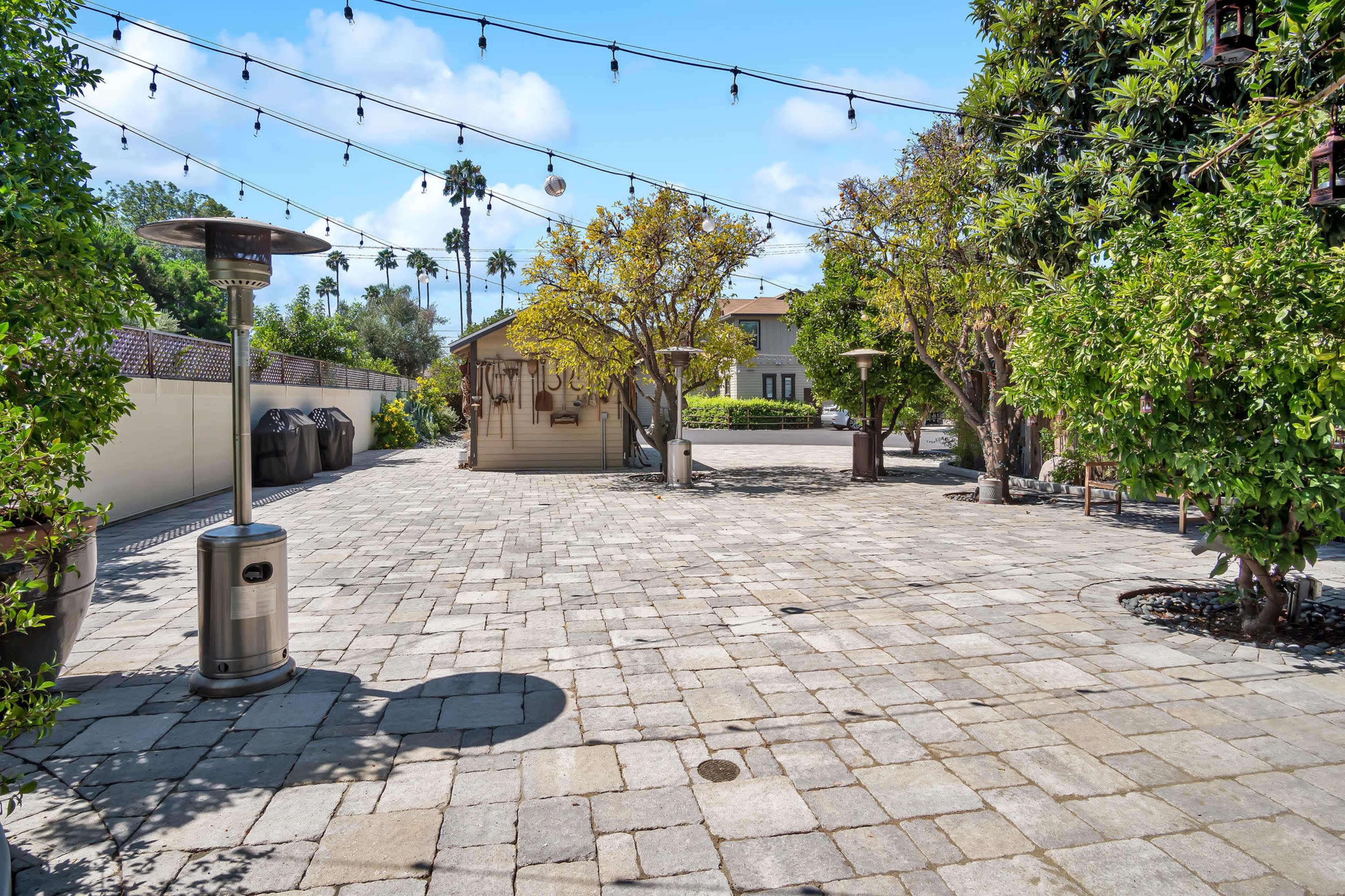 The image shows a landscaped outdoor area with stone paving, surrounded by trees and string lights, featuring heating lamps and a small structure in the background.