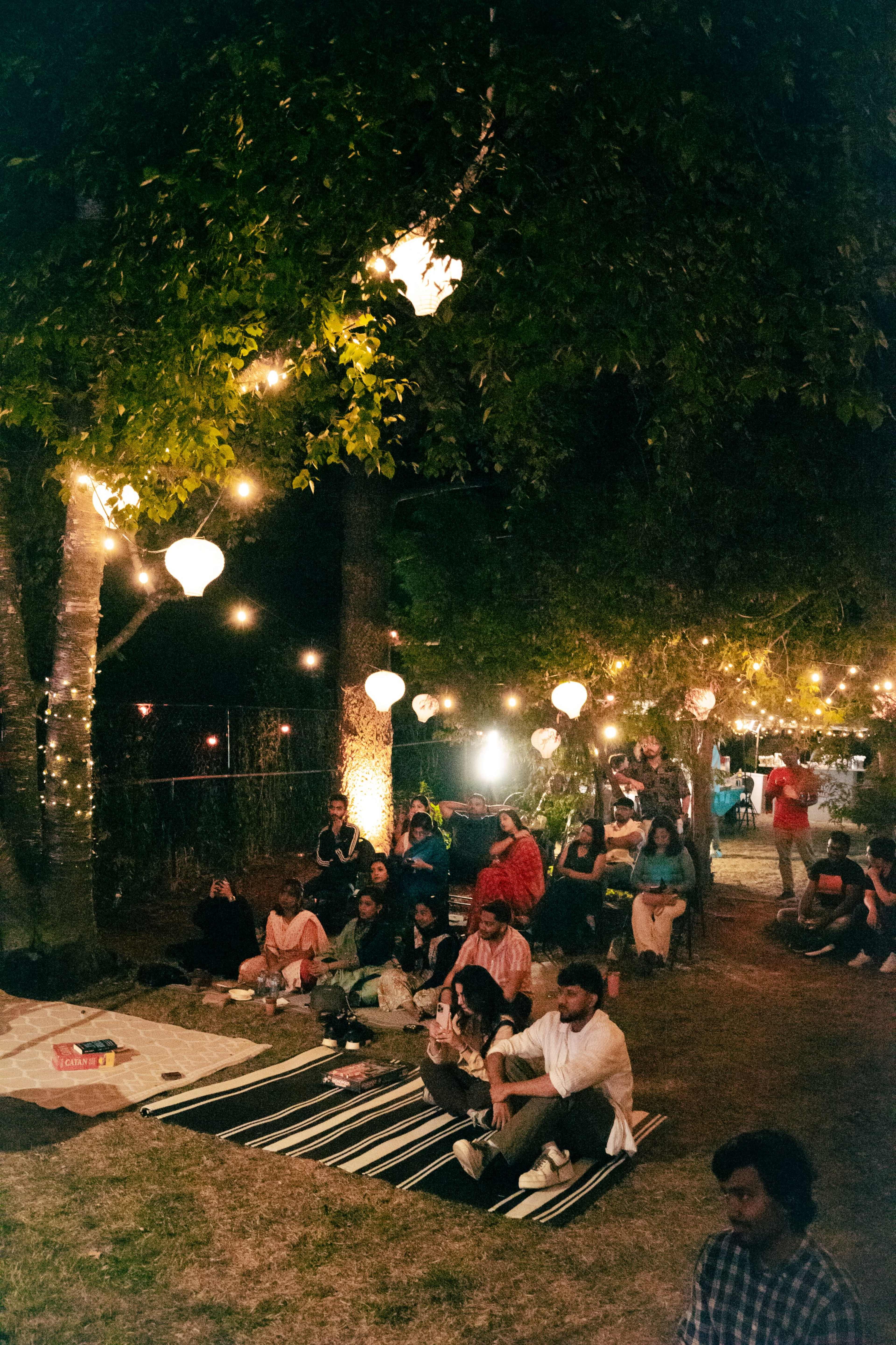 A group of people sits on blankets in a dimly lit outdoor space decorated with hanging lanterns and twinkling lights.
