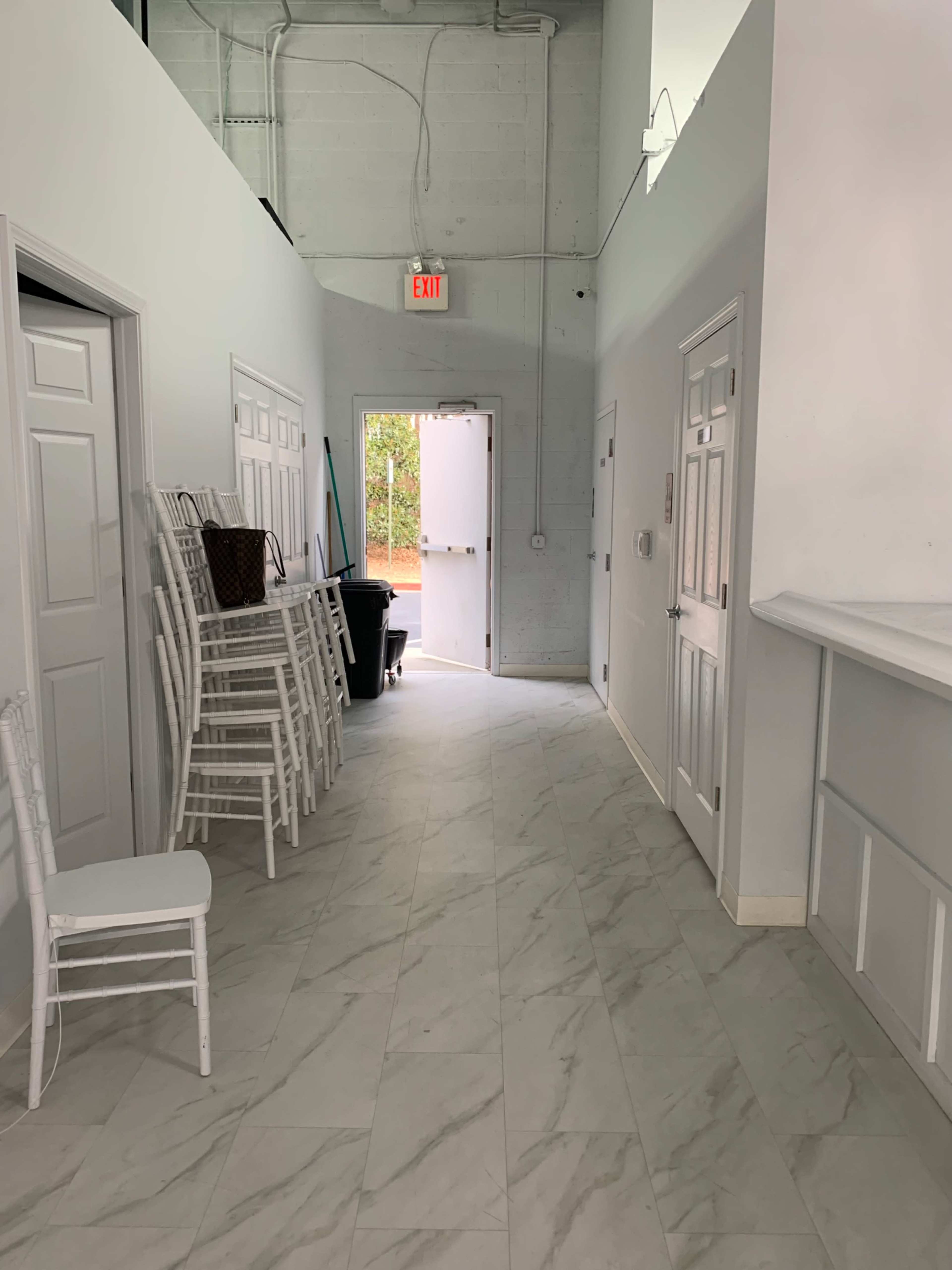 A long hallway with white walls, chairs stacked against one side, and a door leading outside at the end.