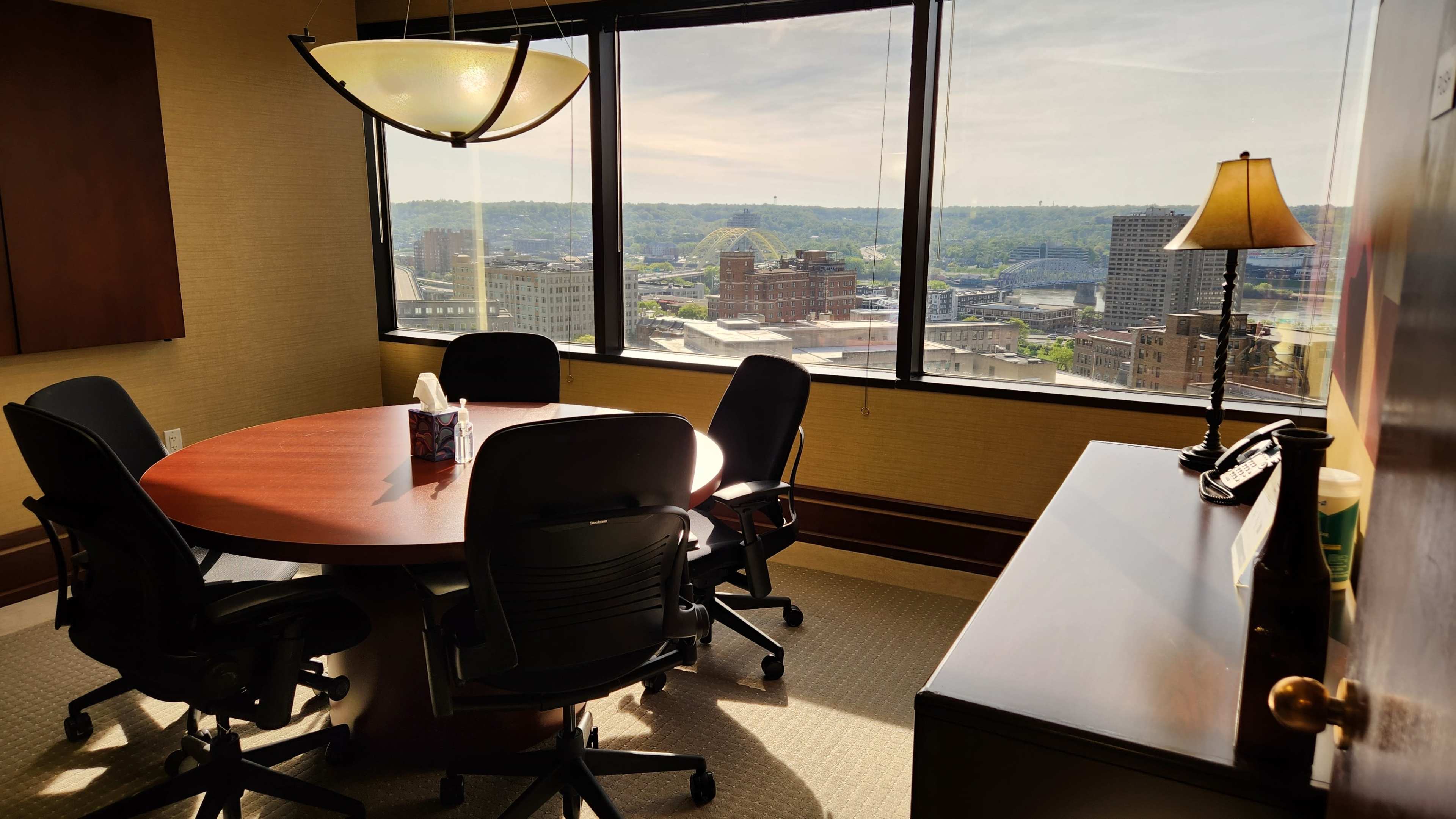 An office meeting room features a round table surrounded by chairs, with a view of city buildings and a bridge visible through the large window.