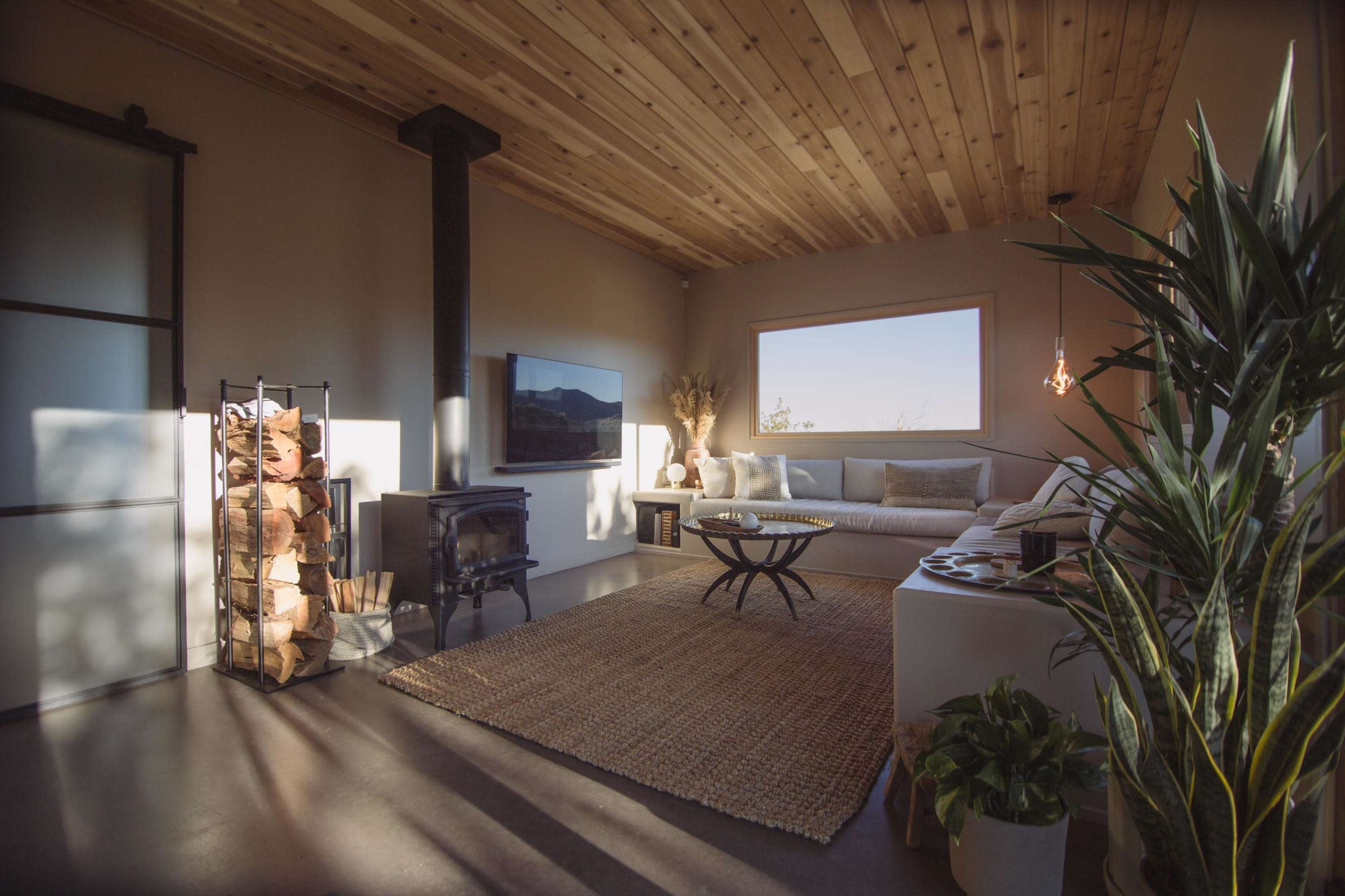 A modern living room features a wooden ceiling, a cozy sofa, a wood stove, and a stack of firewood beside a large window.