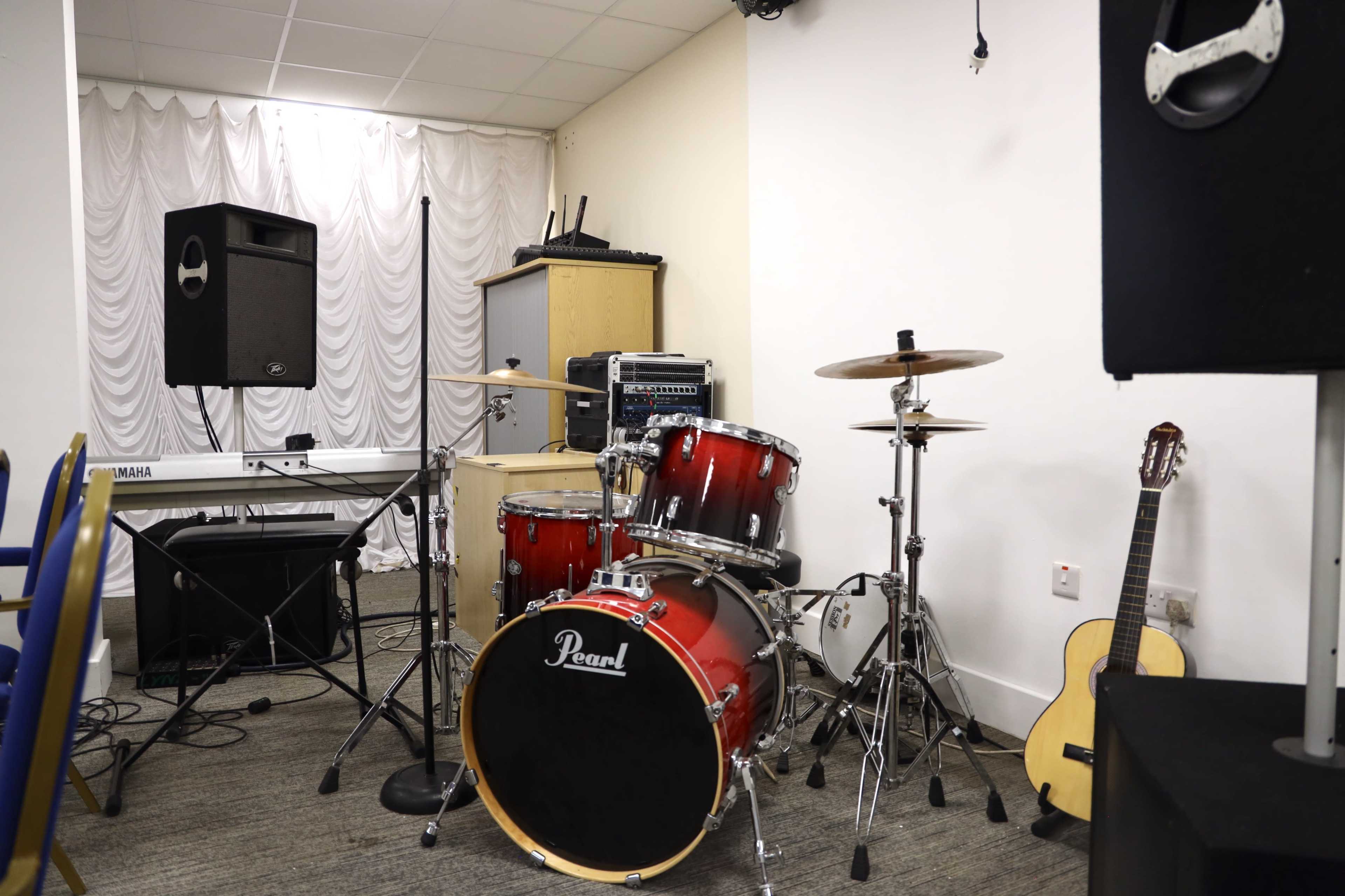 The image shows a small music setup featuring a drum kit, a keyboard, an amplifier, and an acoustic guitar against a wall in a room with white drapery.