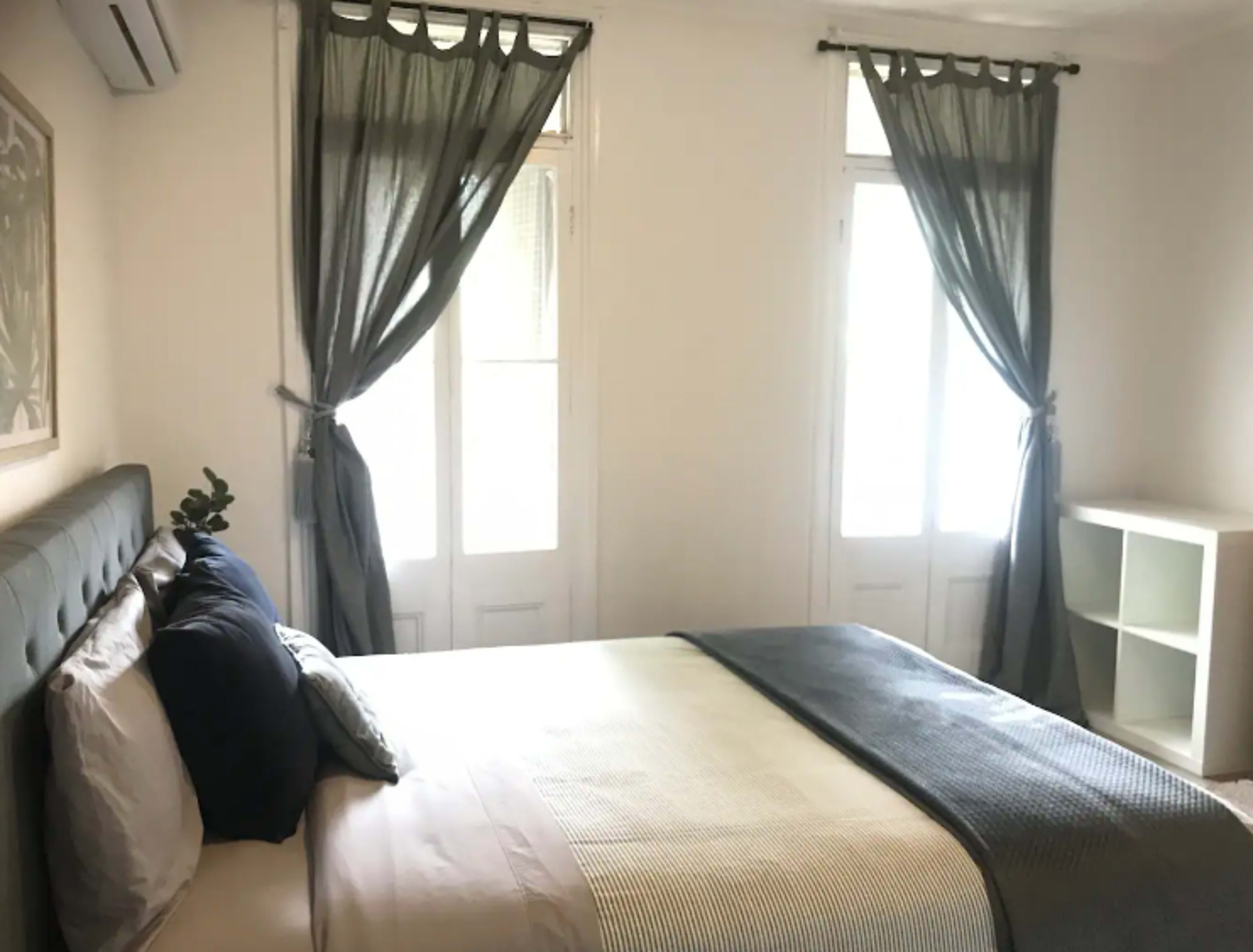 A bedroom with a neatly made bed, two windows dressed with sheer curtains, and a simple white shelf against the wall.