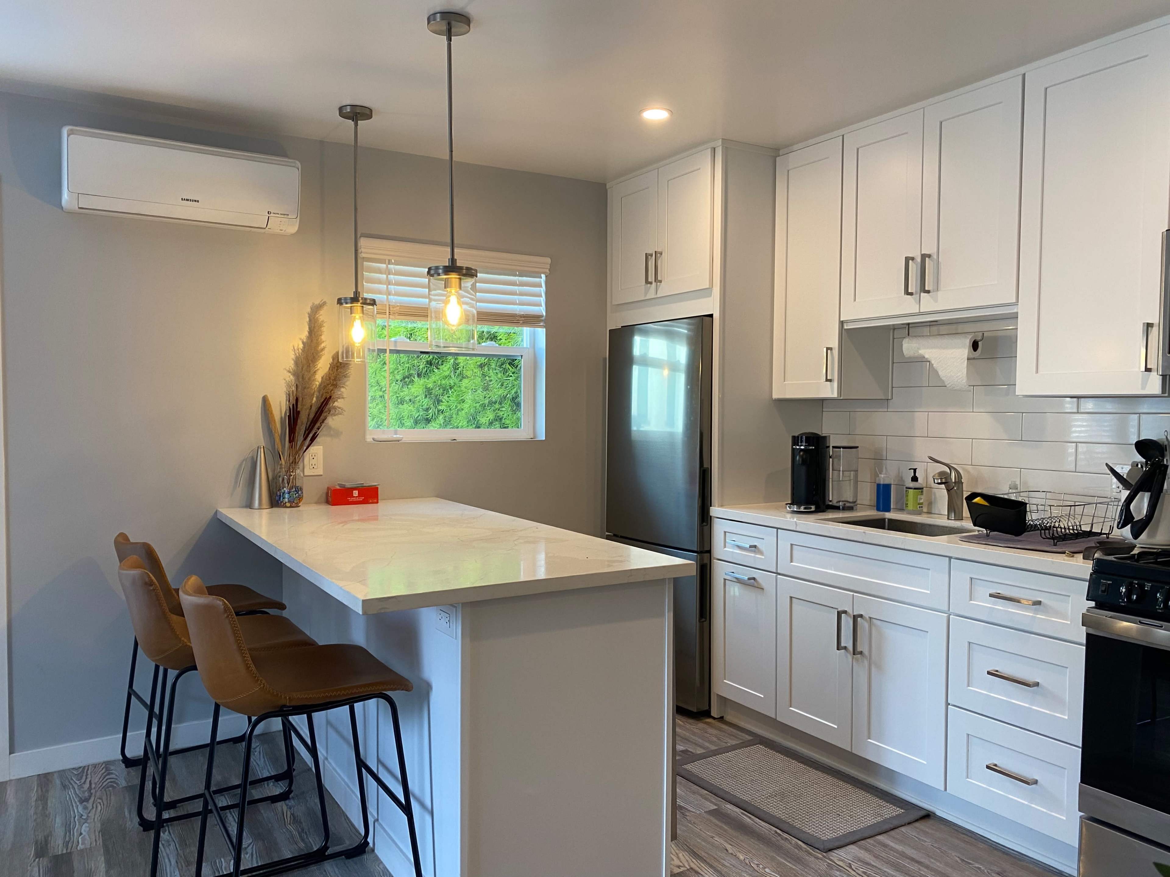 A modern kitchen features white cabinetry, a central island with three bar stools, and natural light coming through a window.