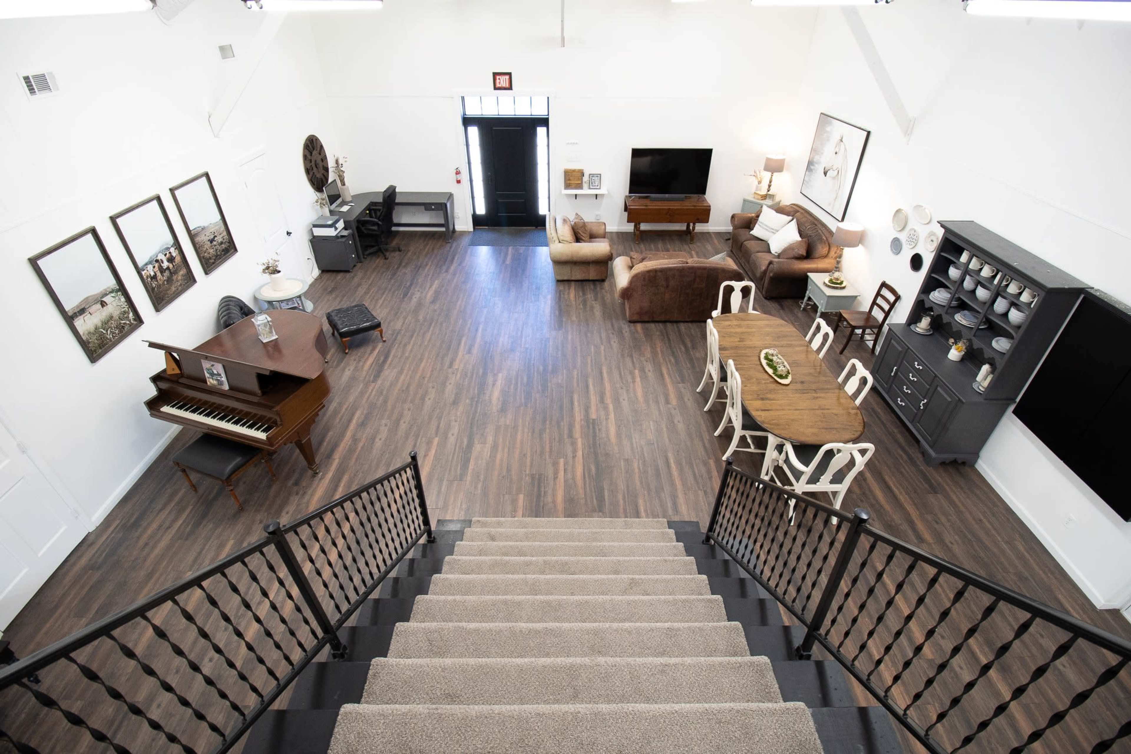 A spacious, modern living area with a staircase leading down to a room featuring a piano, dining table, seating arrangements, and various decorative elements.