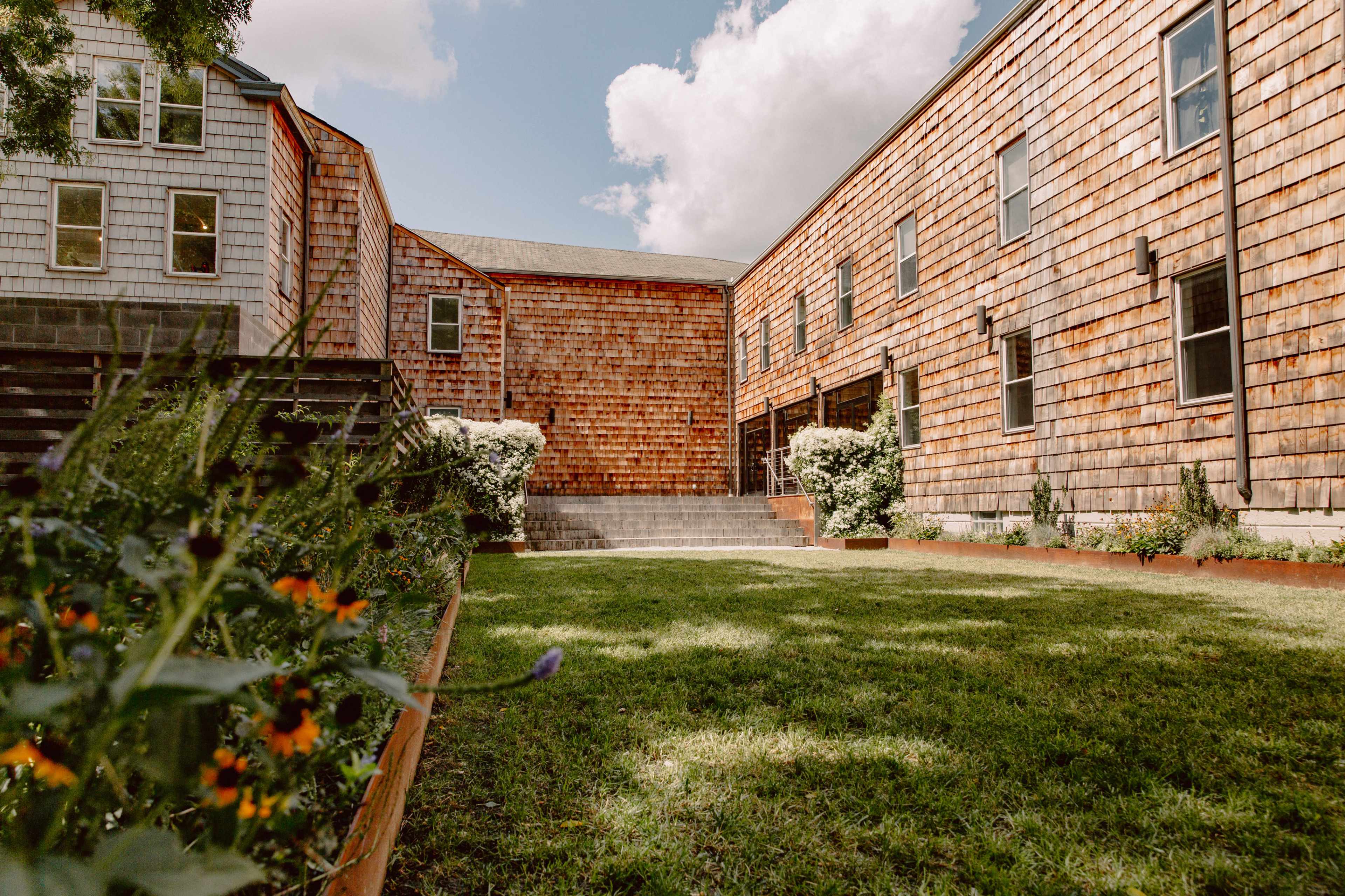 The image shows a courtyard surrounded by wooden buildings, with a grassy area in the center and landscaped flower beds along the edges.