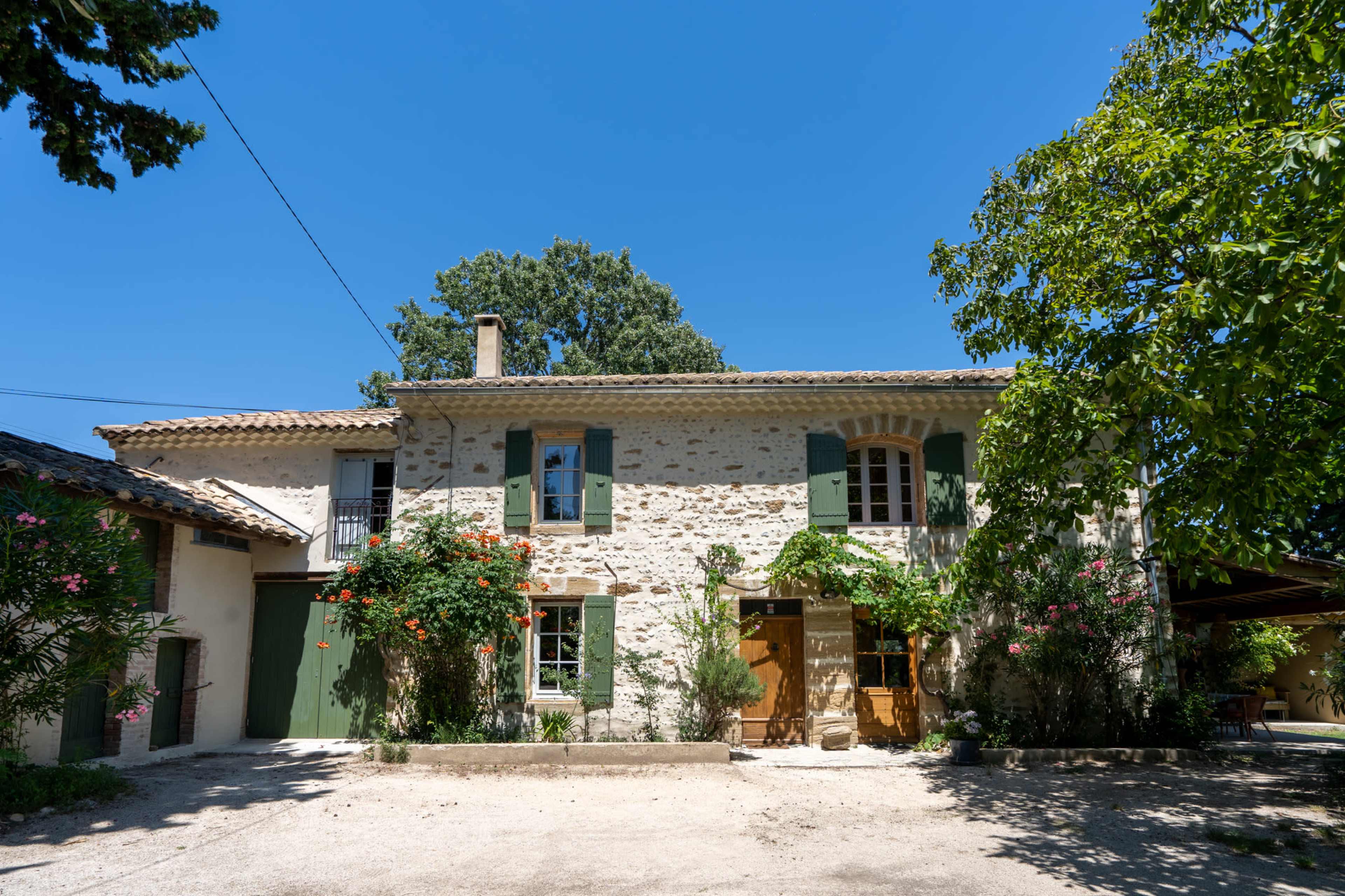 A stone house with green shutters and a wooden door is surrounded by trees and flowering plants under a clear blue sky.