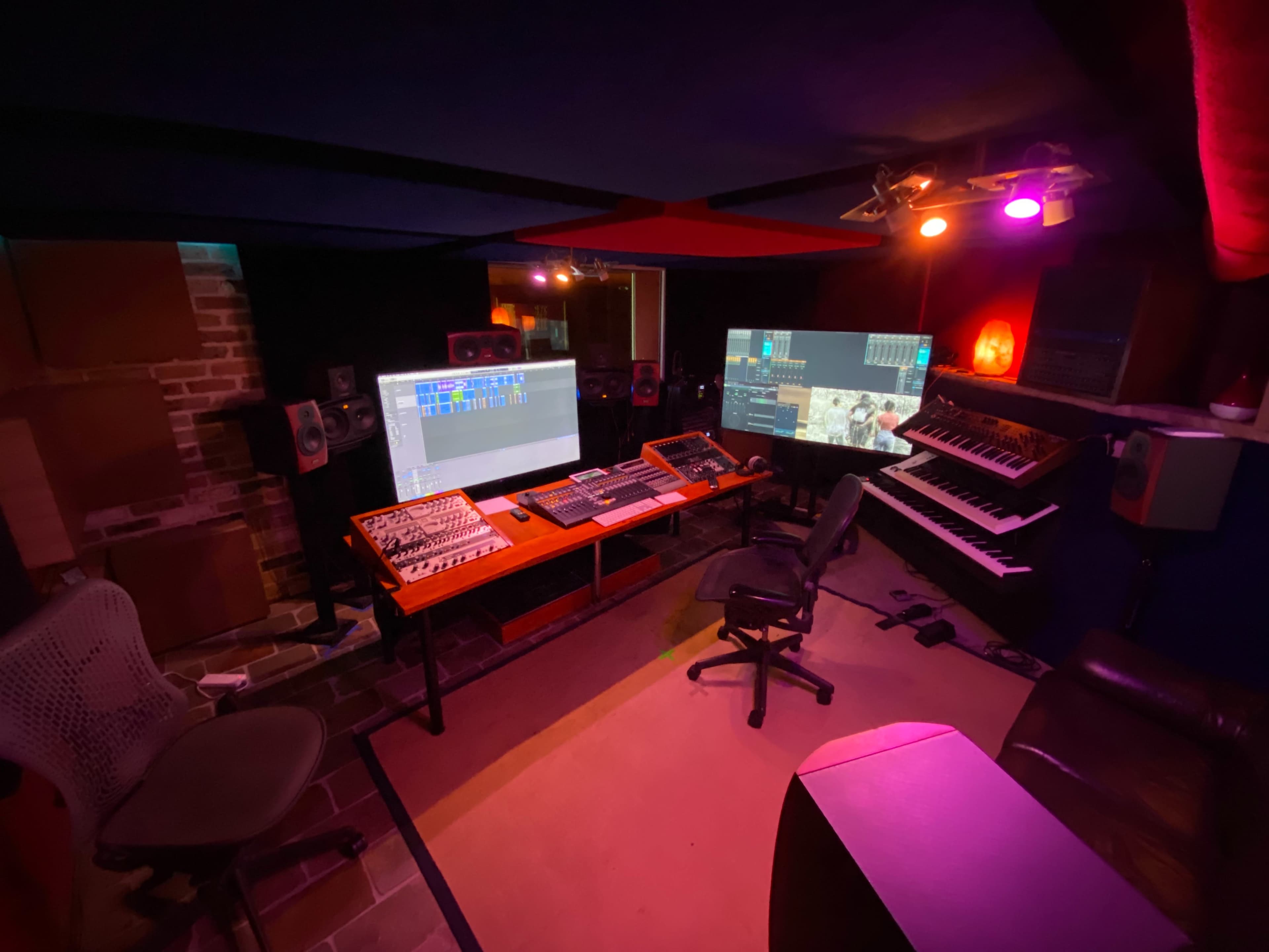 The image shows a music studio with multiple monitors, a keyboard, audio equipment, and seating arranged around a central workstation.