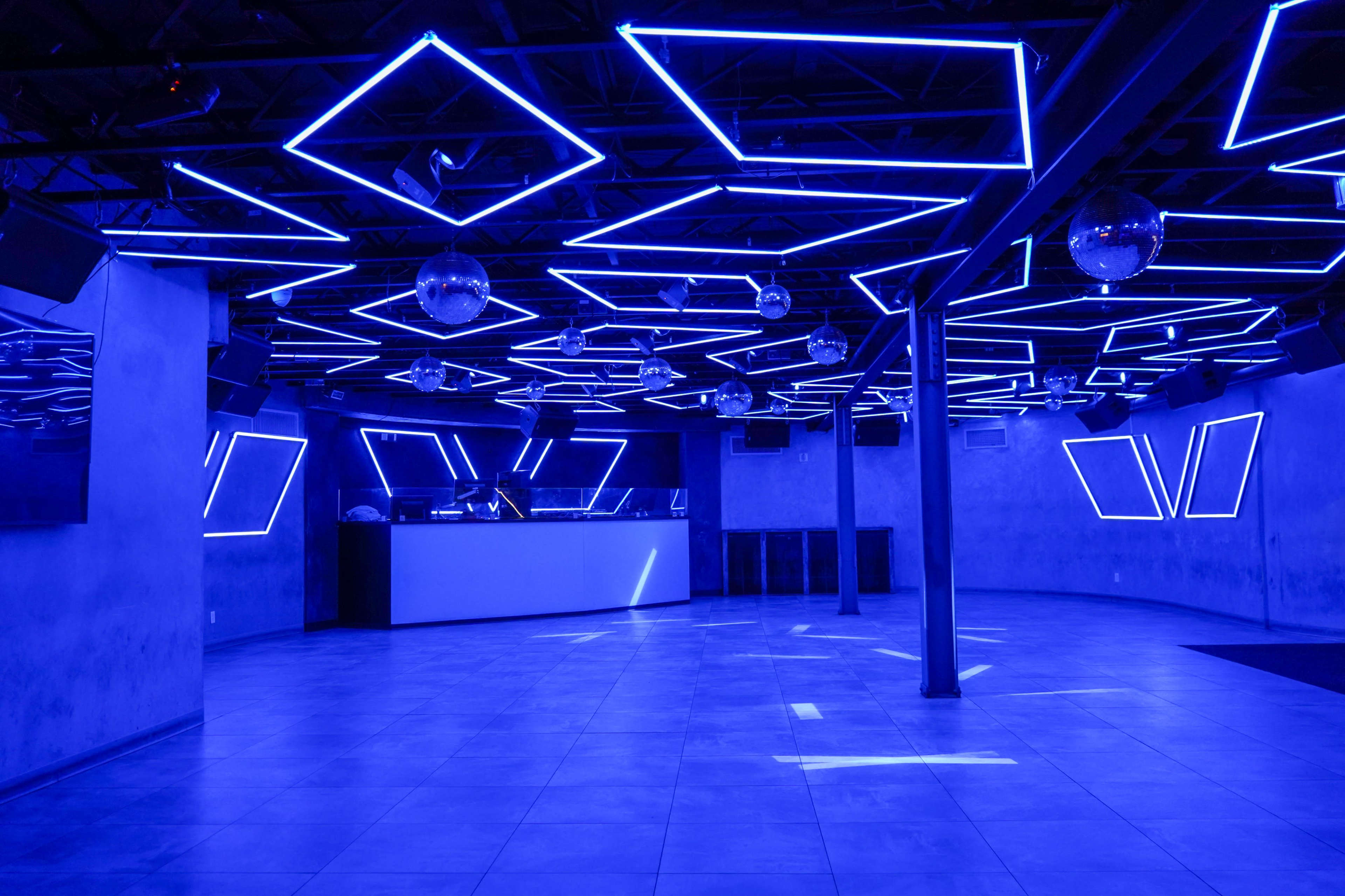 A nightclub interior features blue neon lighting with geometric shapes on the ceiling and a bar area against a wall.