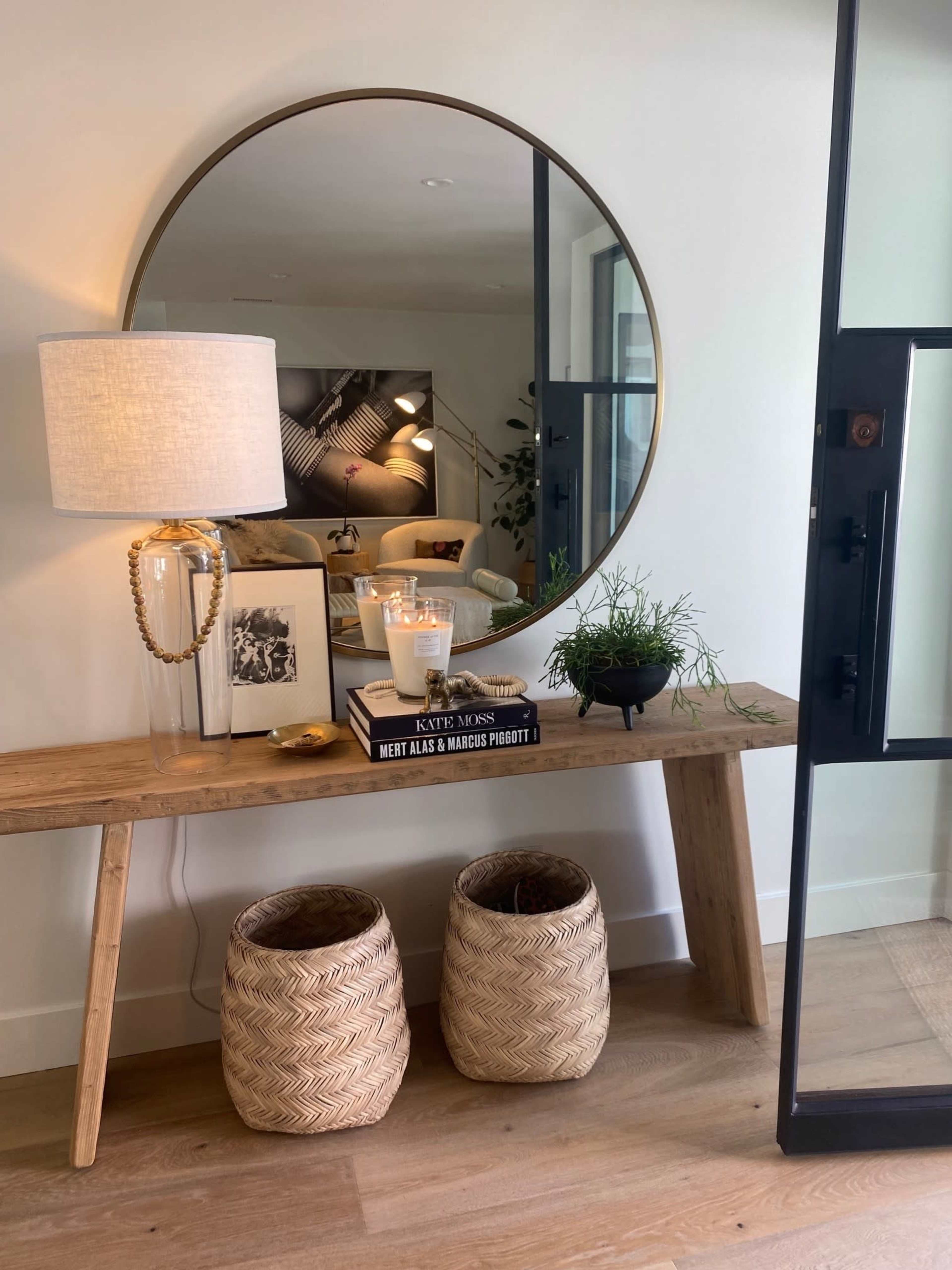 A wooden console table with a round mirror above it features a lamp, decorative items, and two woven baskets underneath.