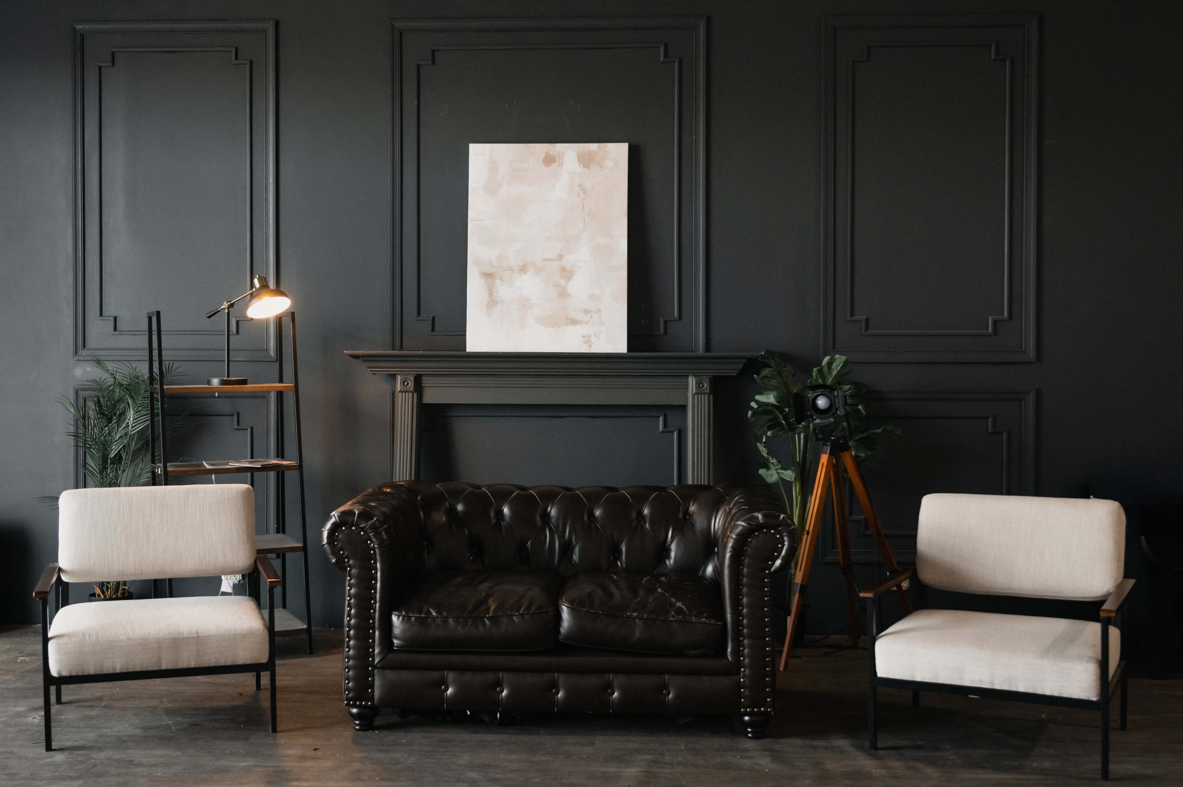 The image features a cozy living room setup with a dark leather chesterfield sofa flanked by two light-colored armchairs, set against a dark wall with a minimalist artwork above a traditional fireplace.