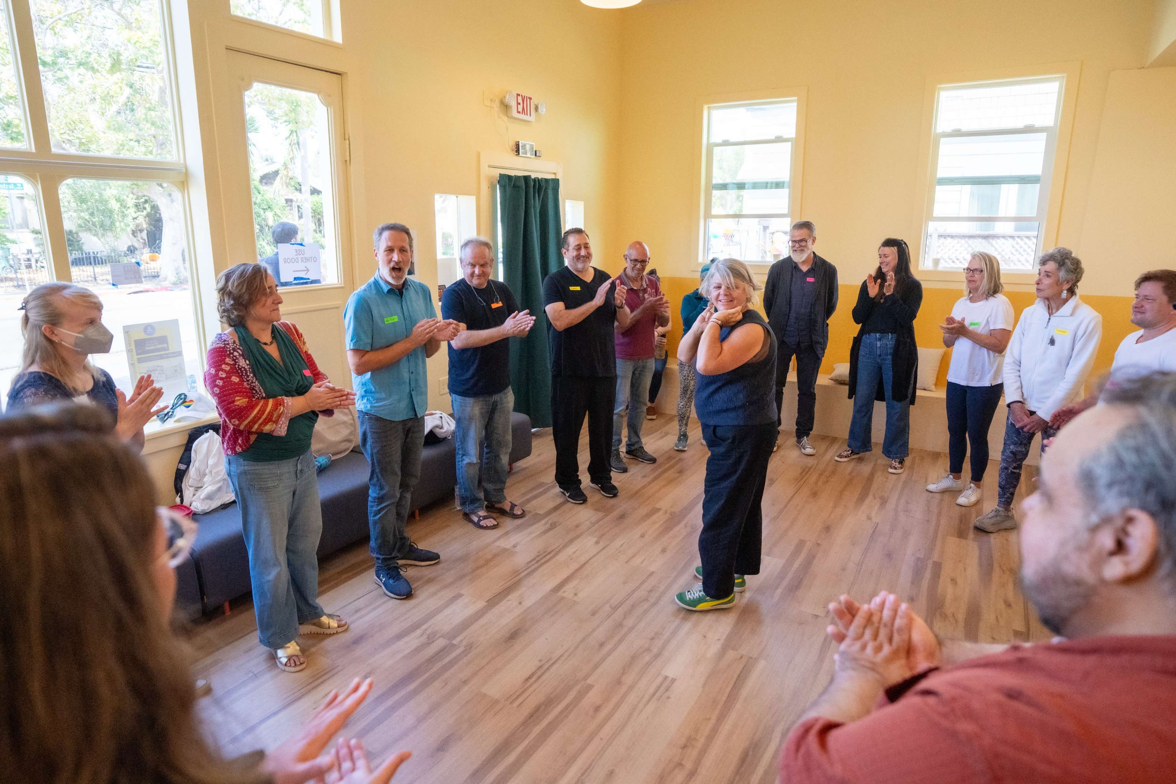 A group of people is standing in a circle, participating in an interactive activity in a warmly lit room with large windows.