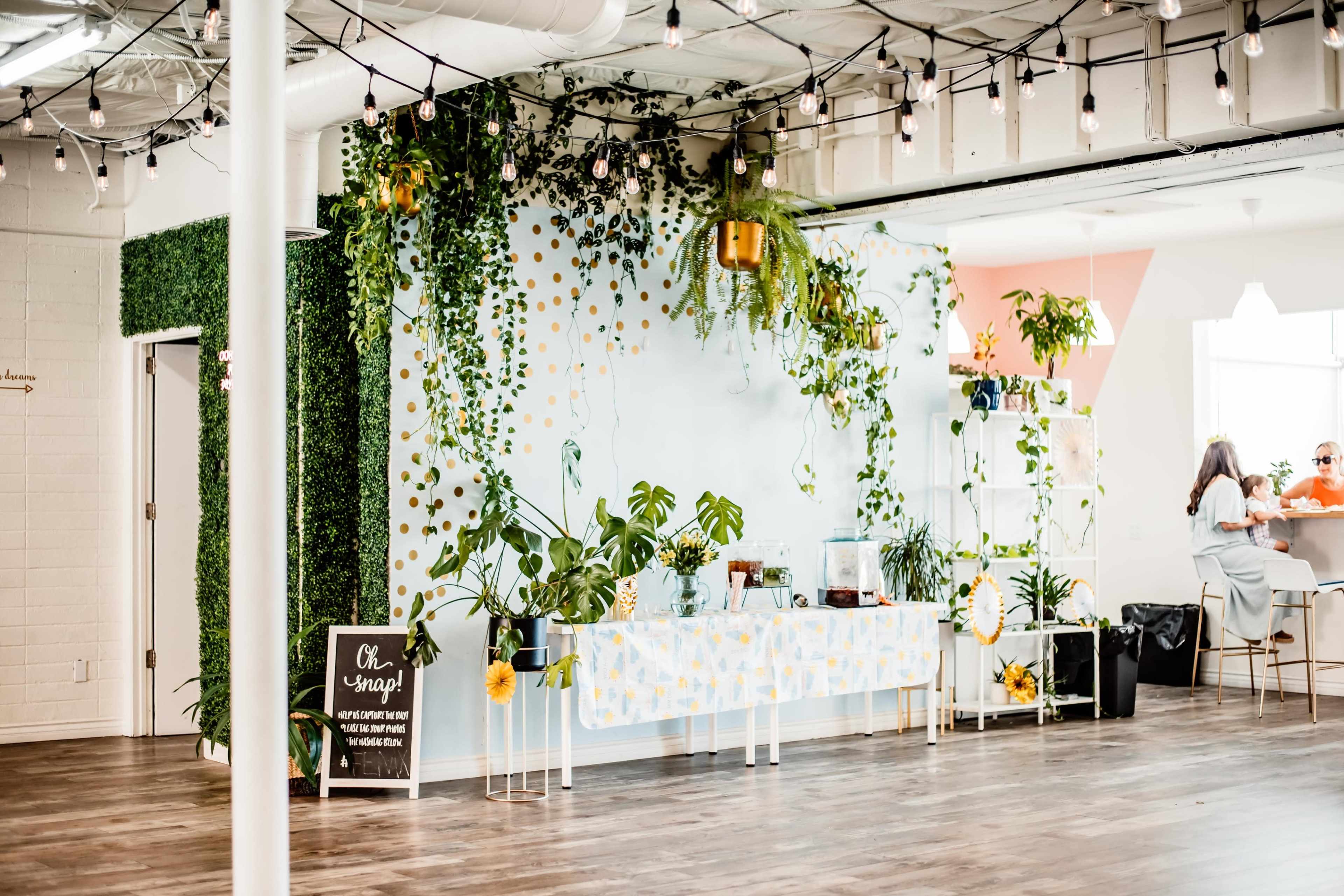 A spacious, well-lit room features a decorated wall with plants and a table set with yellow and white decorations, while people can be seen in the background.