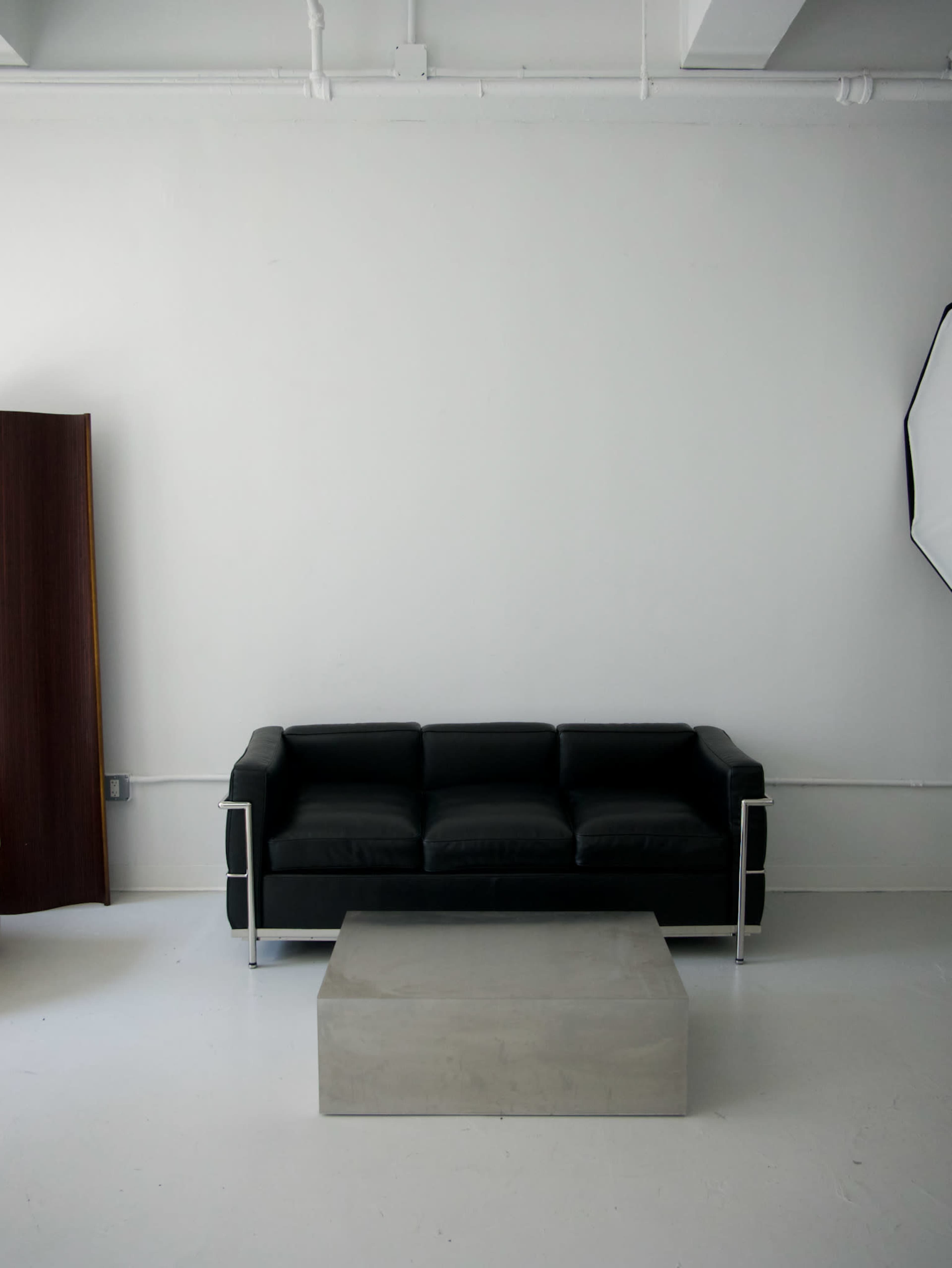 A black sofa with chrome legs is positioned in front of a rectangular concrete coffee table in a minimalist, brightly lit room with white walls.