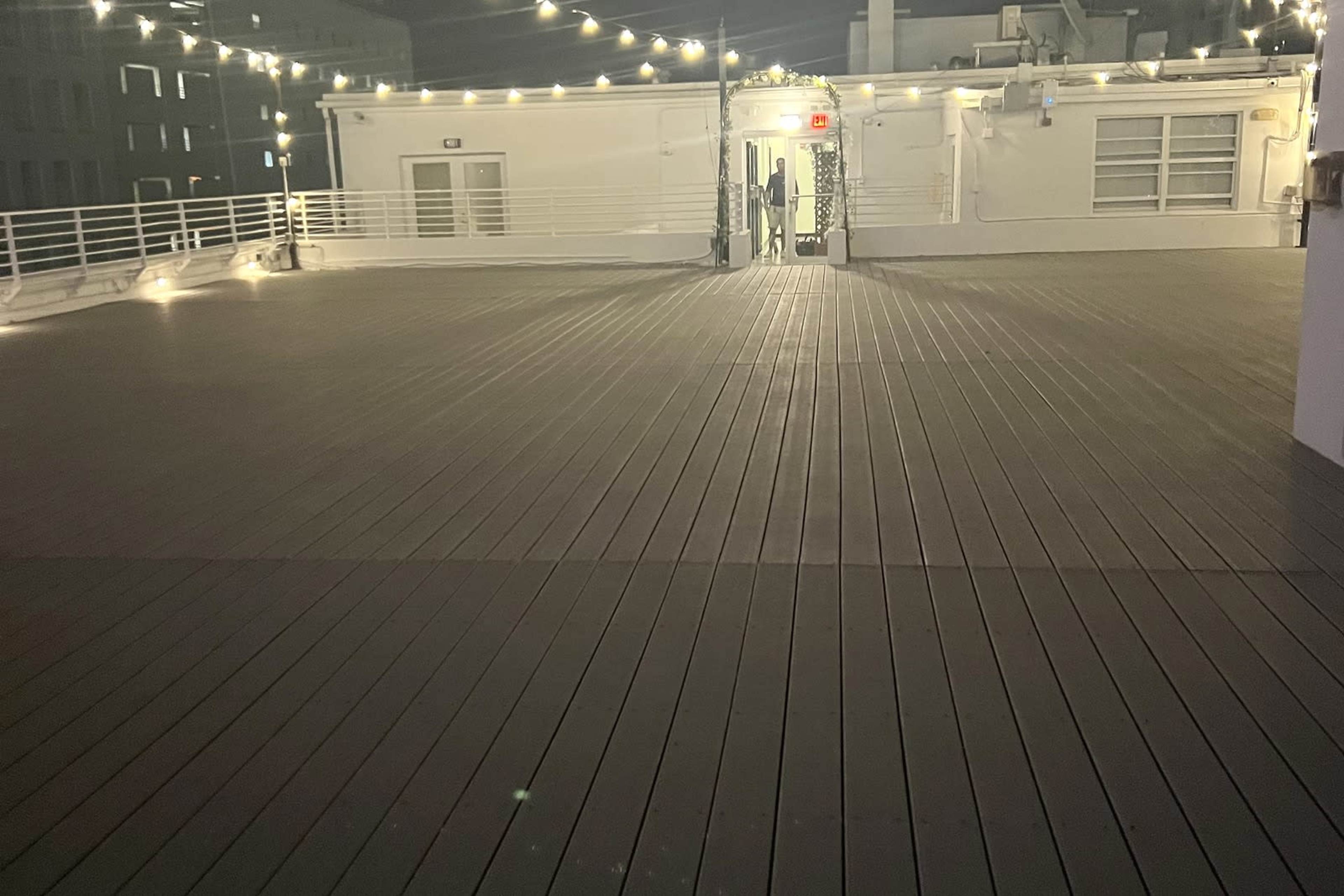 The image shows an empty rooftop deck with wooden flooring and string lights illuminating the area.