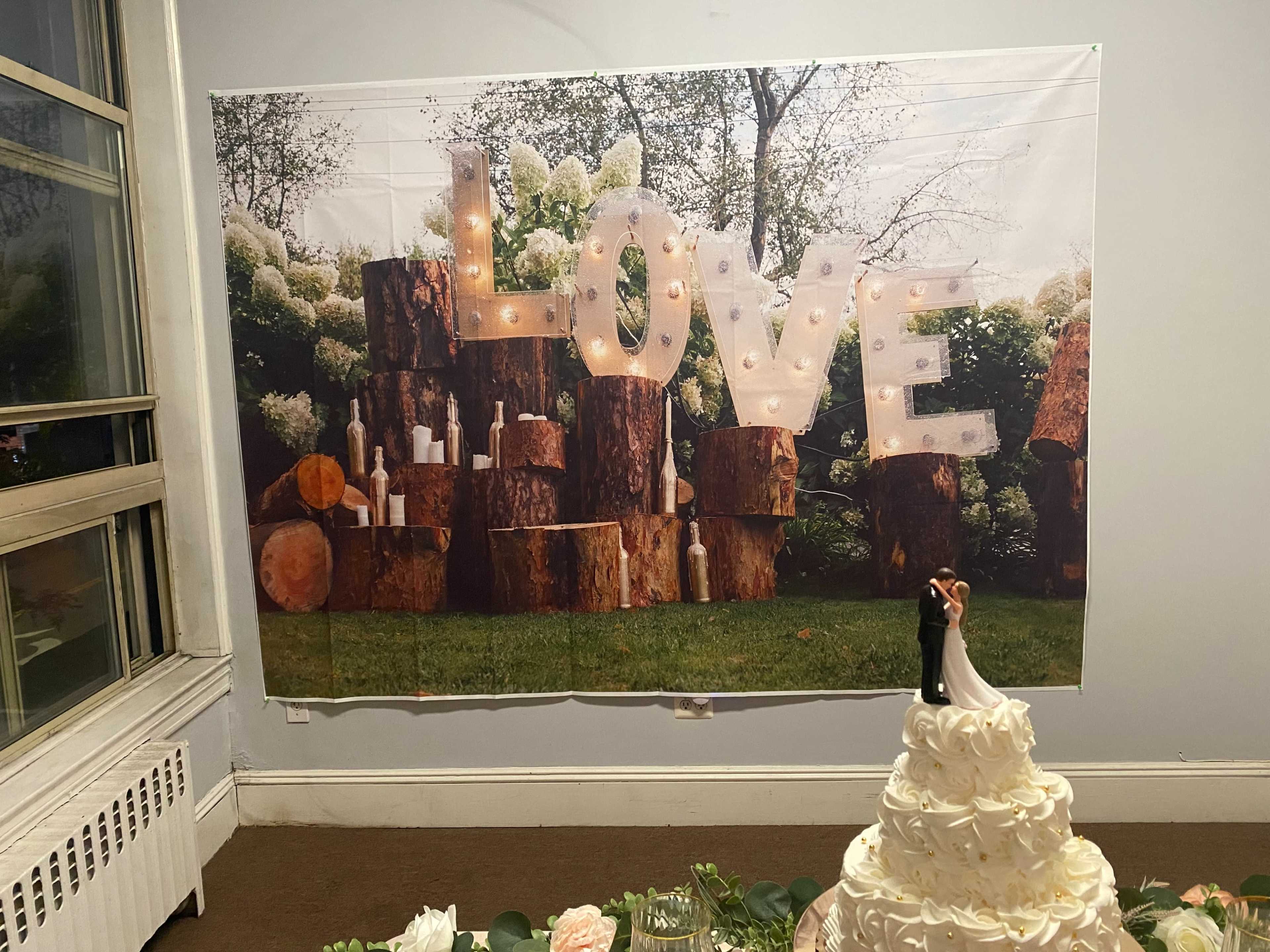 A large backdrop displaying the word "LOVE" in illuminated letters with a scene of tree stumps and greenery is positioned behind a wedding cake on a table.