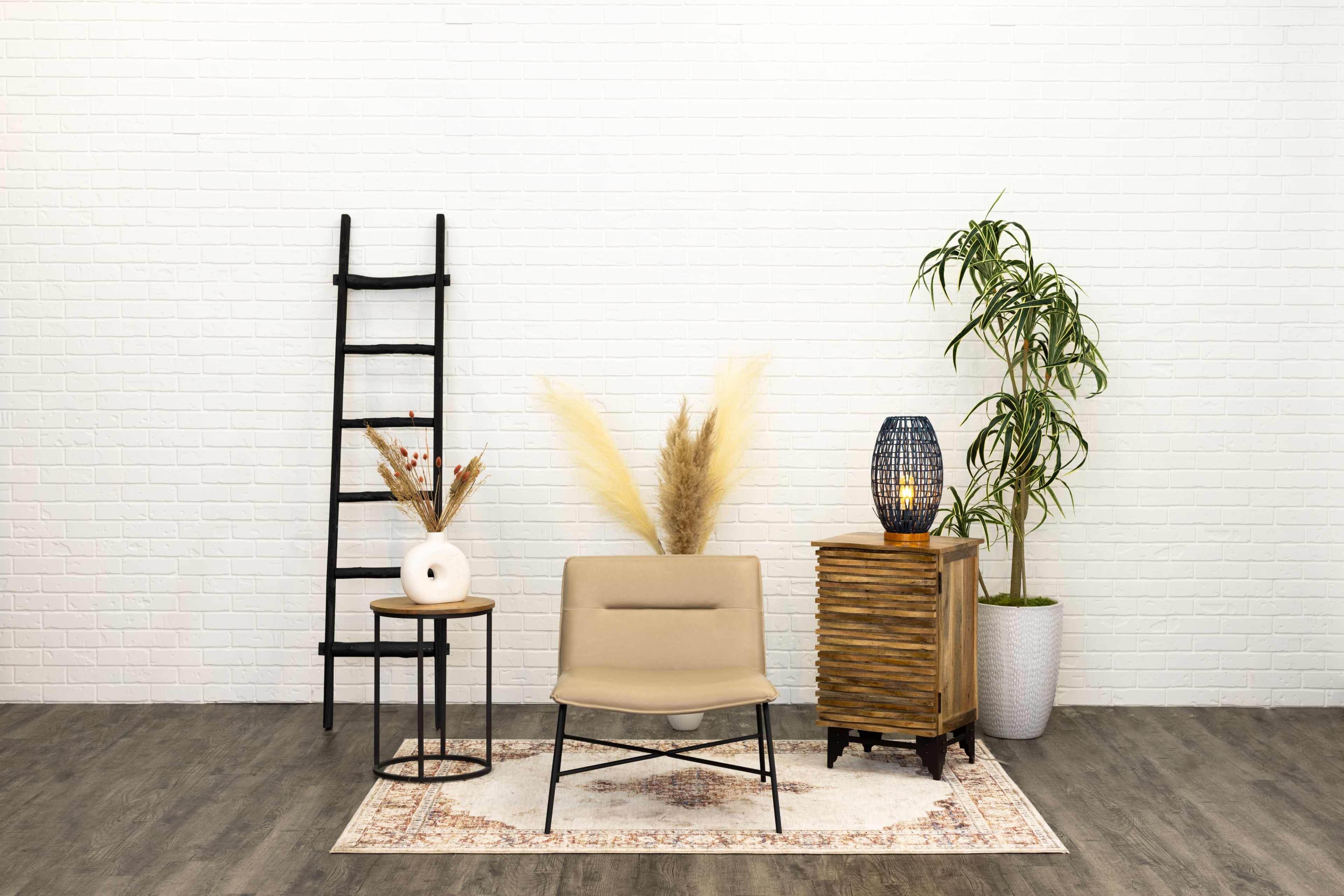 A minimalist interior scene with a beige chair, a decorative ladder, a small side table, a potted plant, and a lamp, all arranged on a patterned rug against a white brick wall.