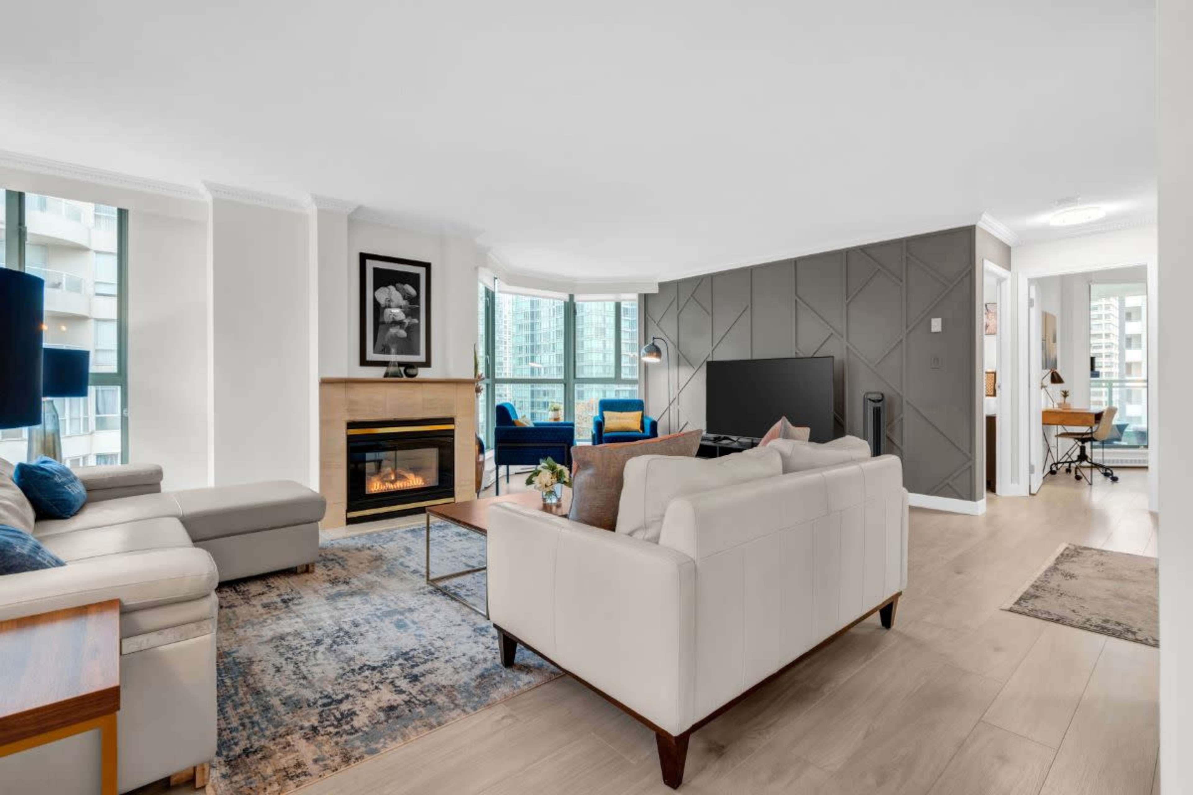 The image shows a modern living room with a fireplace, a large TV, and a mix of seating areas, including a light-colored sofa and an accent chair, set against large windows.